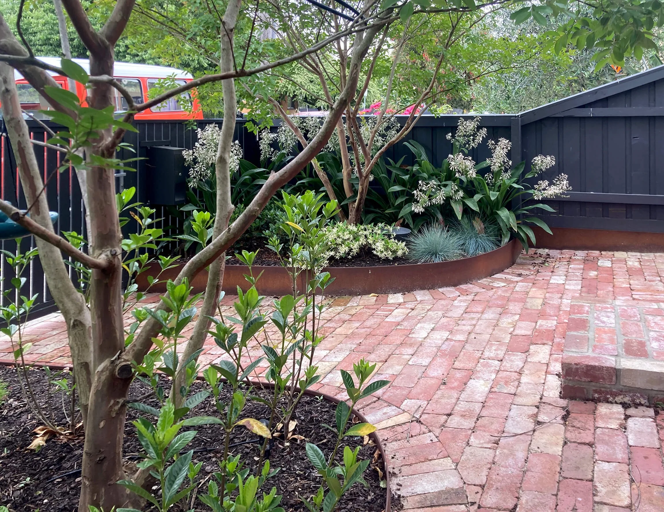 A garden with a brick patio, lush green plants, and trees. A black wooden fence surrounds the garden, and there is a red vehicle partially visible in the background.
