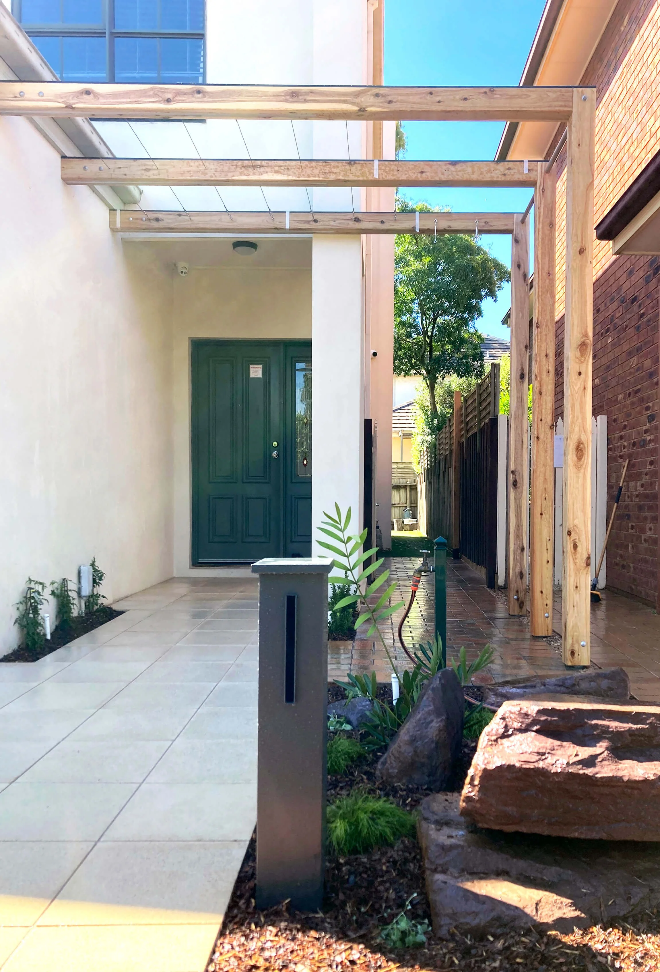 Front yard under construction with wooden framework, potted plants, rocks, a green front door, tiled porch, and a clear blue sky.