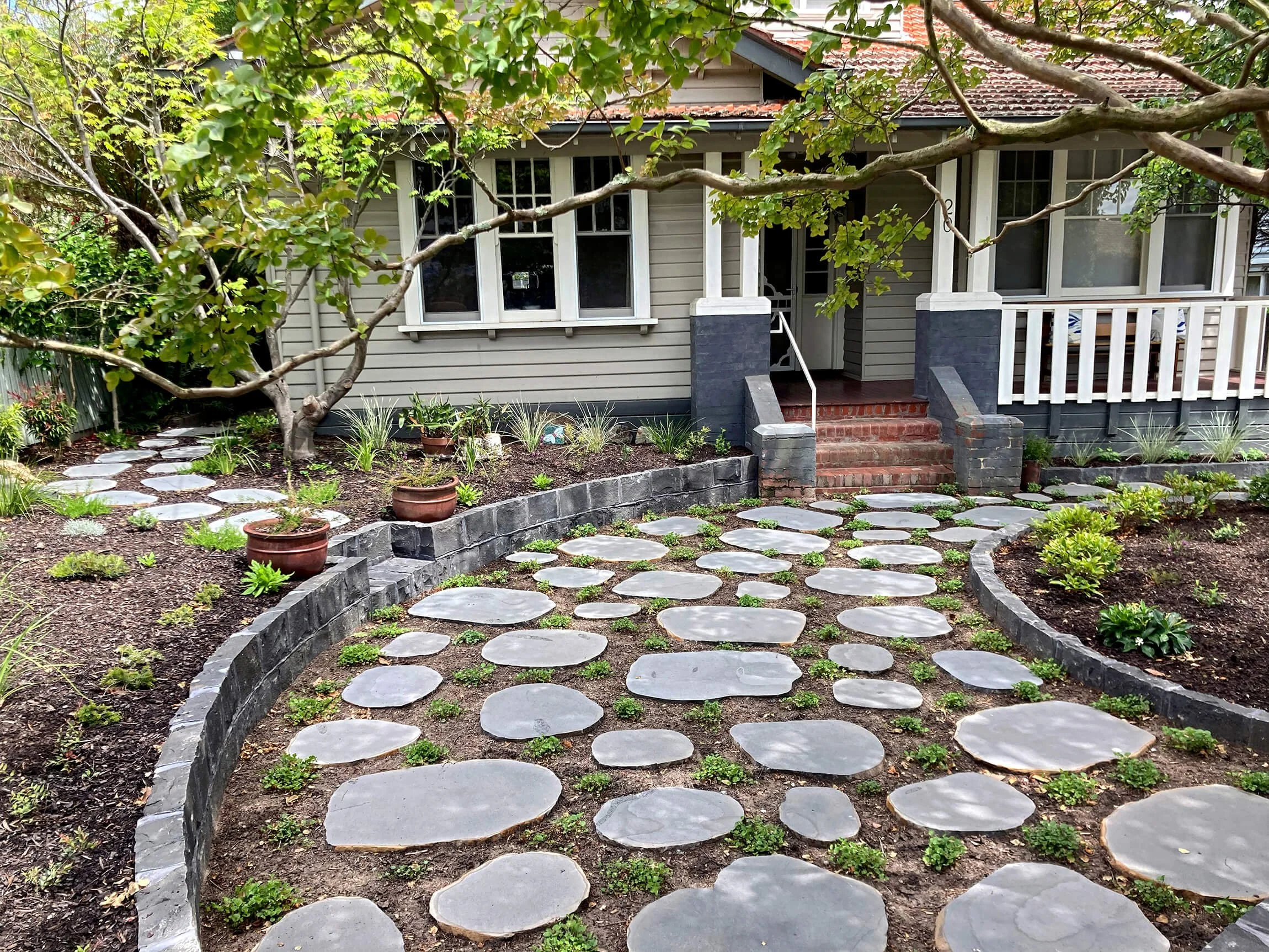 A front yard with a stone pathway leading to a porch of a house. There is a tree with green leaves in the yard, potted plants, and a garden bed with small plants and flowers.