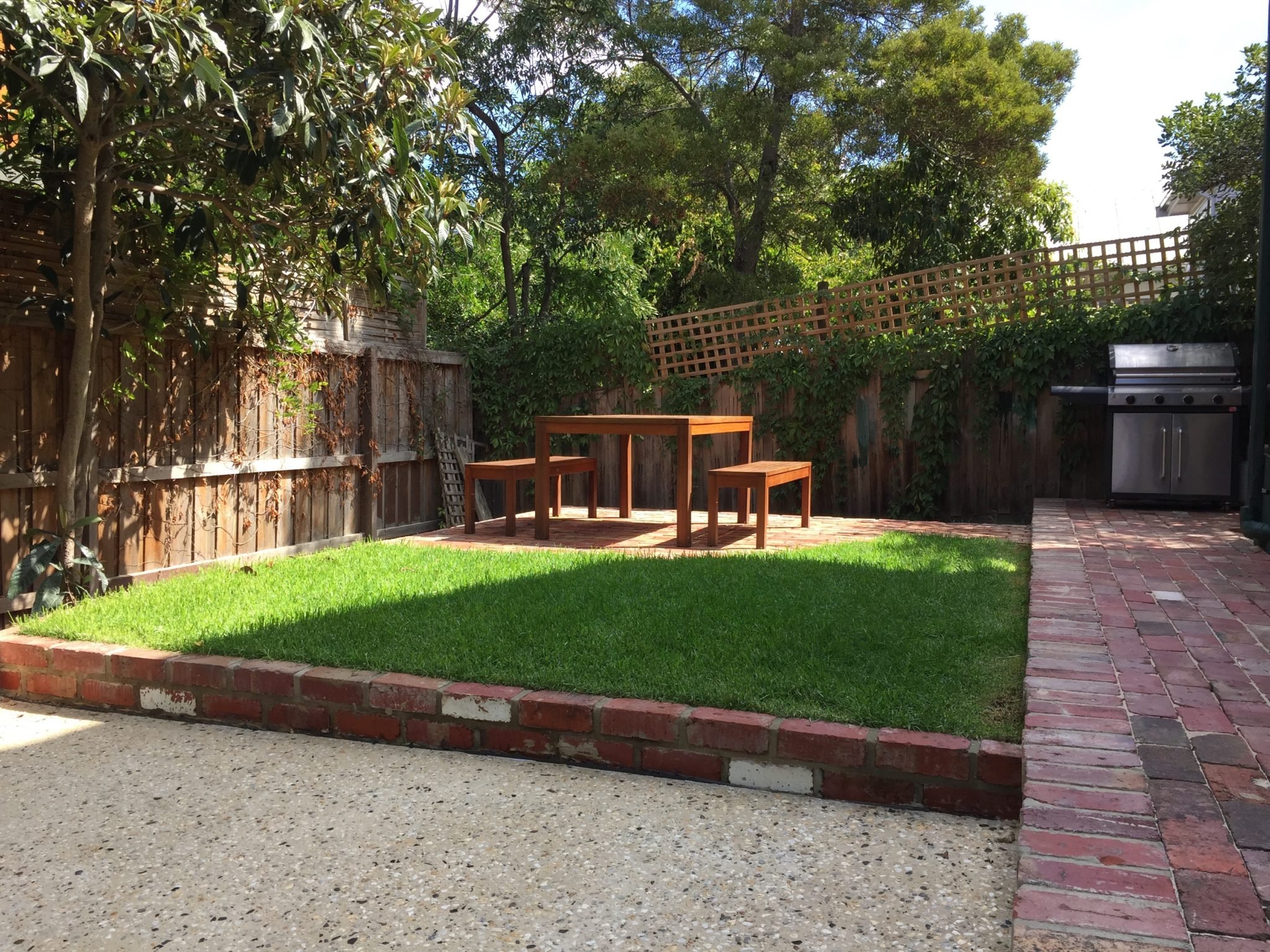 A backyard patio with a wooden table and chairs, a barbecue grill, a brick pathway, a grassy lawn, and a wooden fence surrounded by trees and shrubbery.
