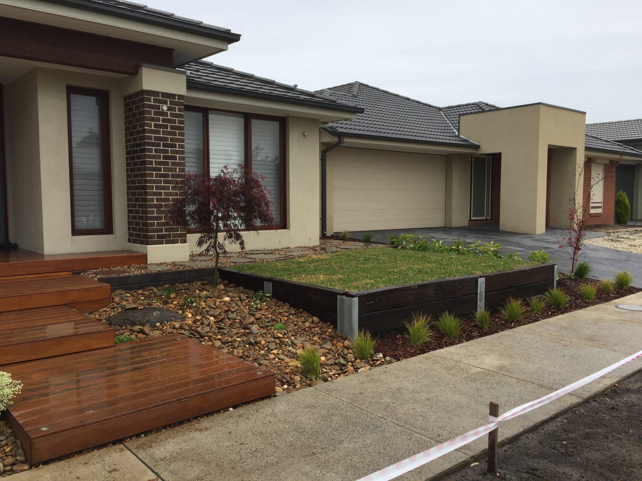 Front yard of a modern house with a concrete driveway, landscaped garden beds, small trees, and a wooden deck.