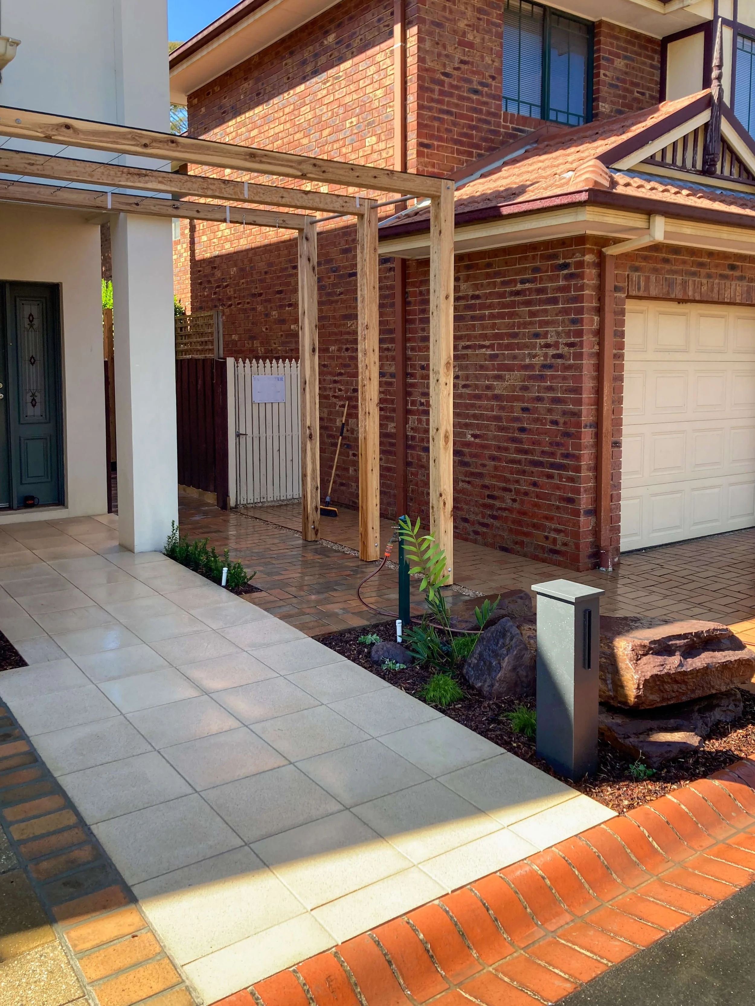 A house exterior with a combination of brick and painted wall, including a garage door, a small garden area with plants, and a partially built wooden pergola structure.