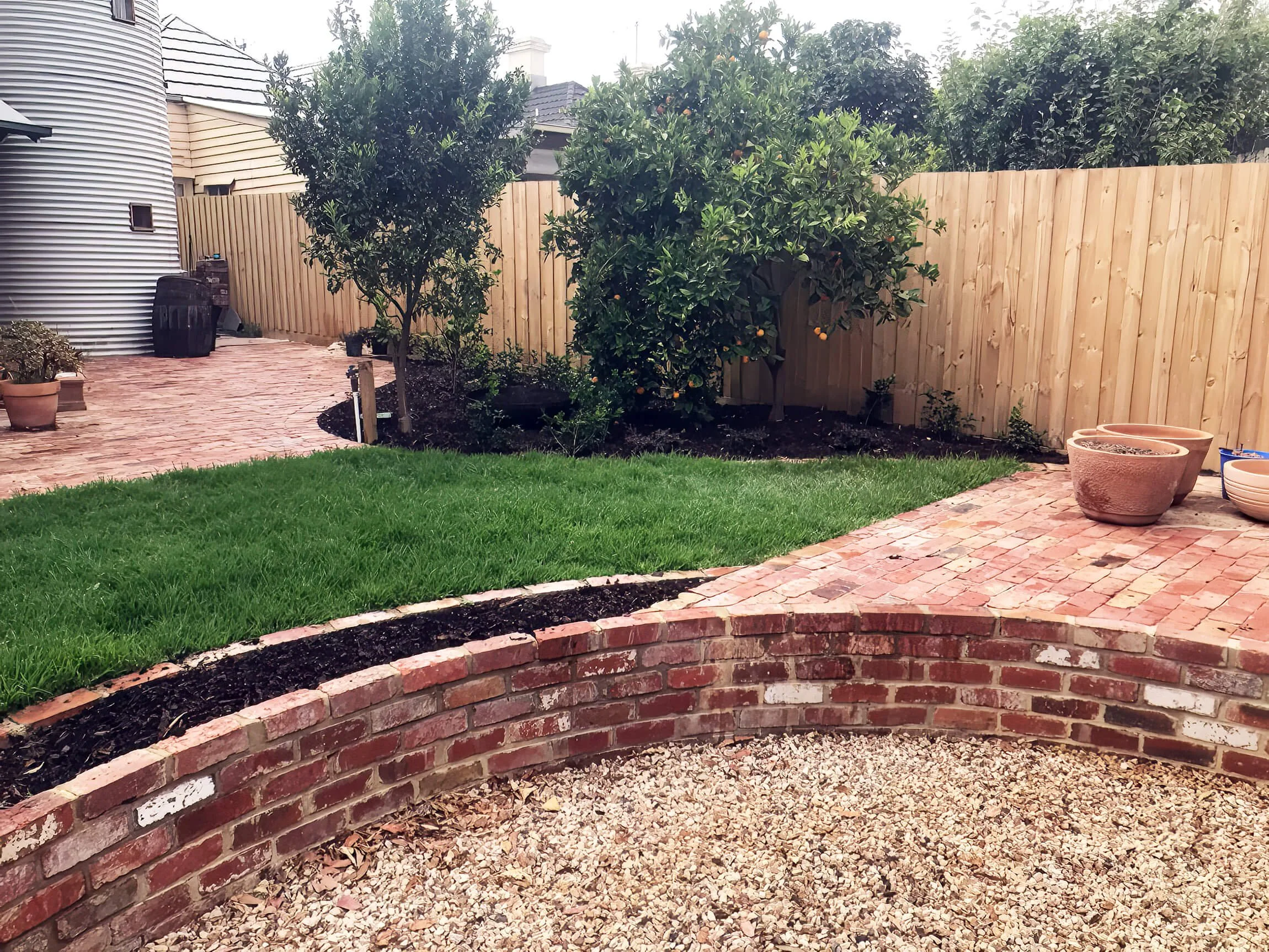 A backyard with a wooden fence, two trees with orange fruit, a brick planter, potted plants, and a brick pathway with grass.