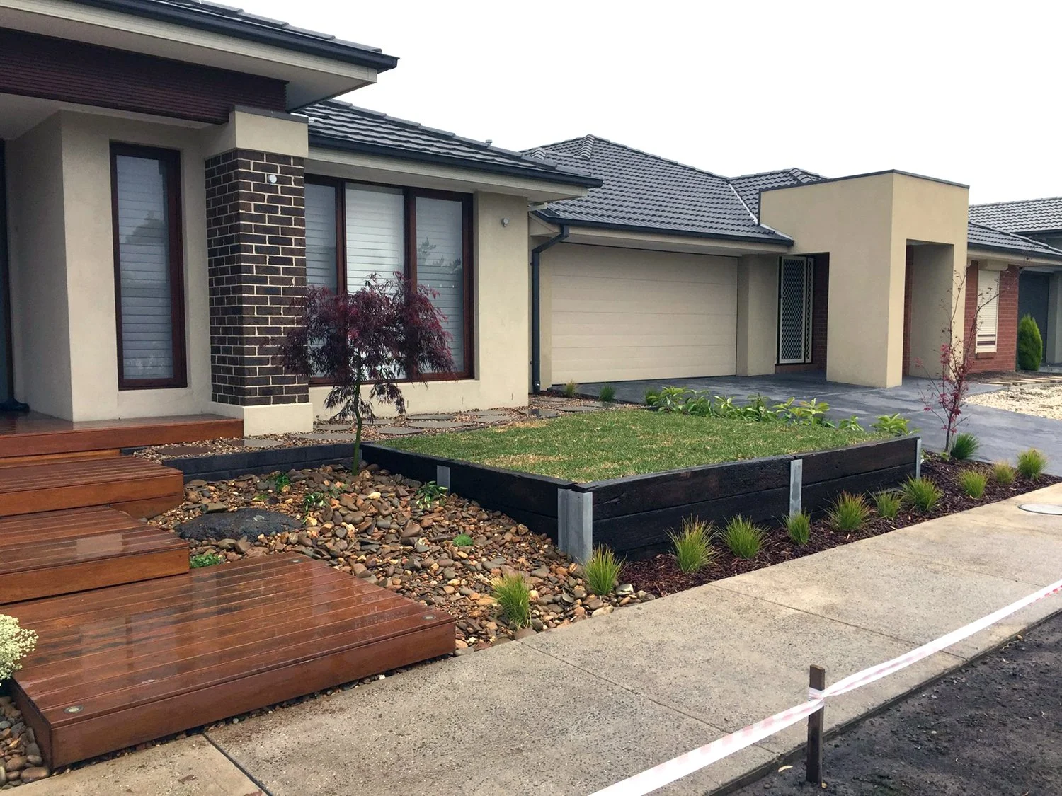 Front yard of a house with wooden steps, a small tree, and a lawn section bordered by black wood. There are some plants along the edge of the yard and a paved driveway leading to a garage. The house has beige walls, brown window frames, and a dark tiled roof.