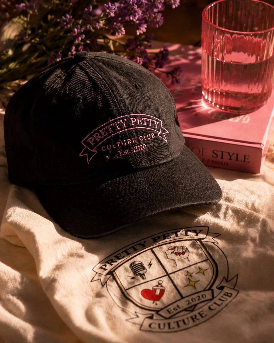 A black cap with pink embroidery reading 'Prettty Petty Culture Club Est. 2020,' placed on a white cloth with a similar logo. In the background, there is a pink book titled 'Of Style' and a pink glass of water on top of a pink napkin, with purple flowers nearby.