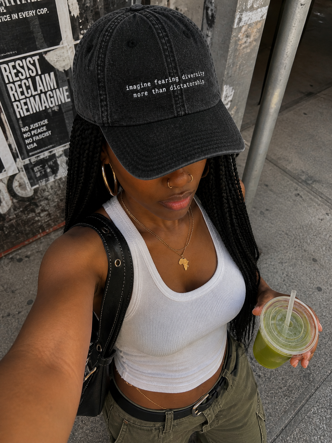 A woman taking a selfie on a city sidewalk, wearing a black baseball cap with the phrase "imagine fearing diversity more than dictatorship," a white tank top, green cargo pants, gold jewelry, and holding a green drink.