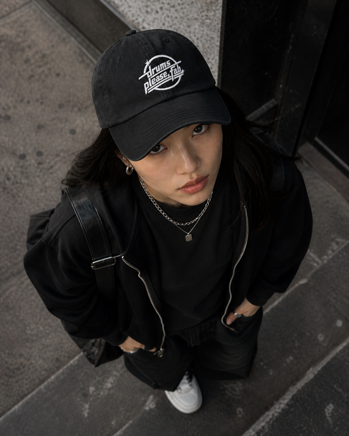 A young woman with dark hair and light brown eyes is looking up at the camera. She is wearing a black cap with white text, a black jacket, layered necklaces, and white sneakers, standing on a concrete sidewalk.