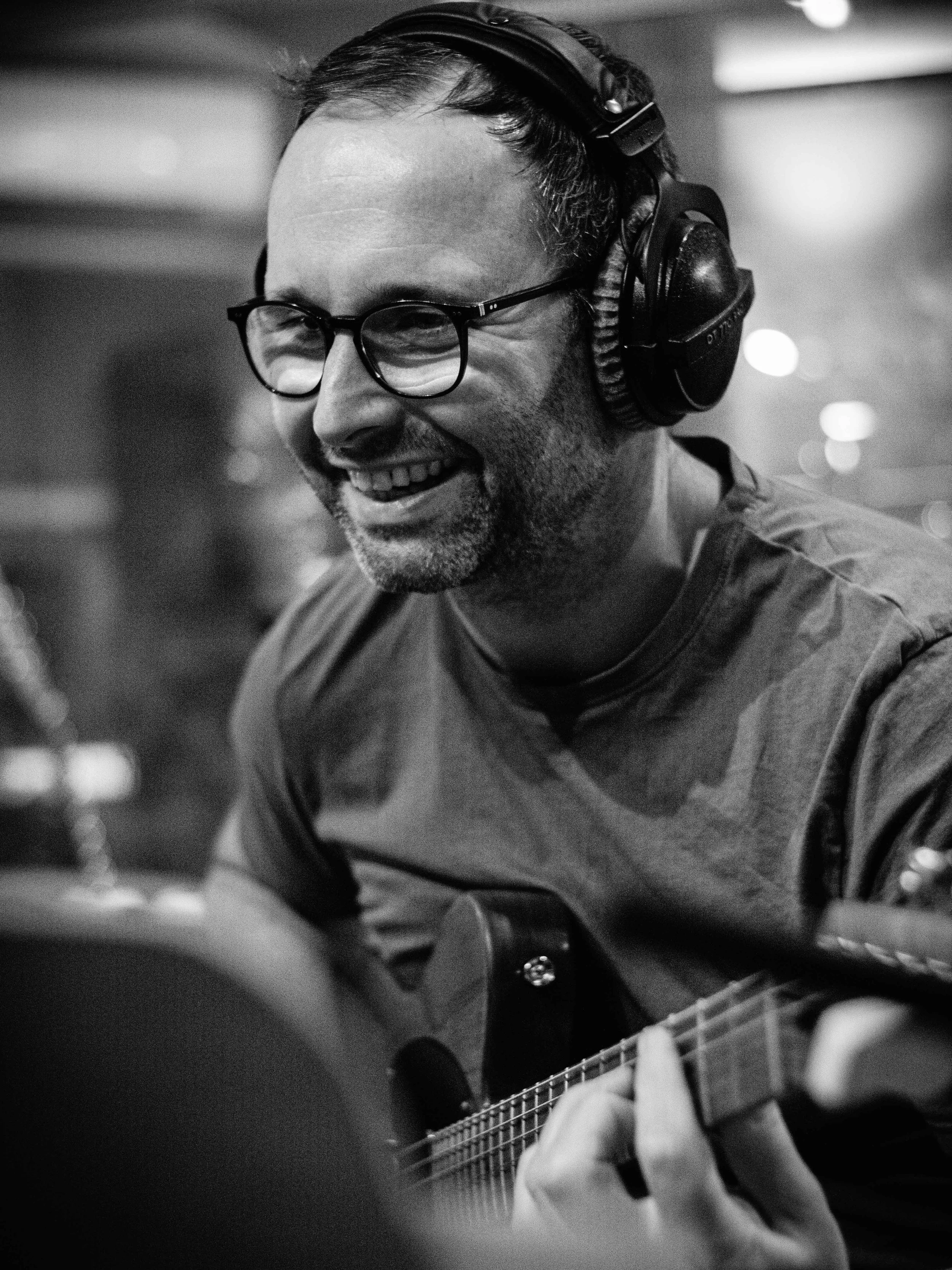 Man wearing glasses and headphones playing an acoustic guitar and smiling.