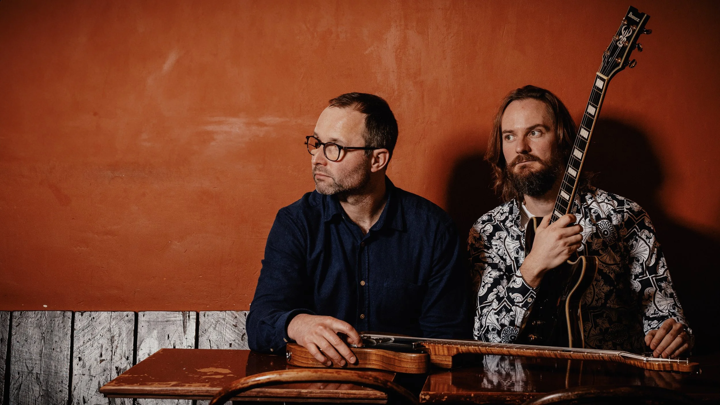 Two men sitting at a wooden table against an orange wall, one holding a guitar and the other with a guitar case on the table.