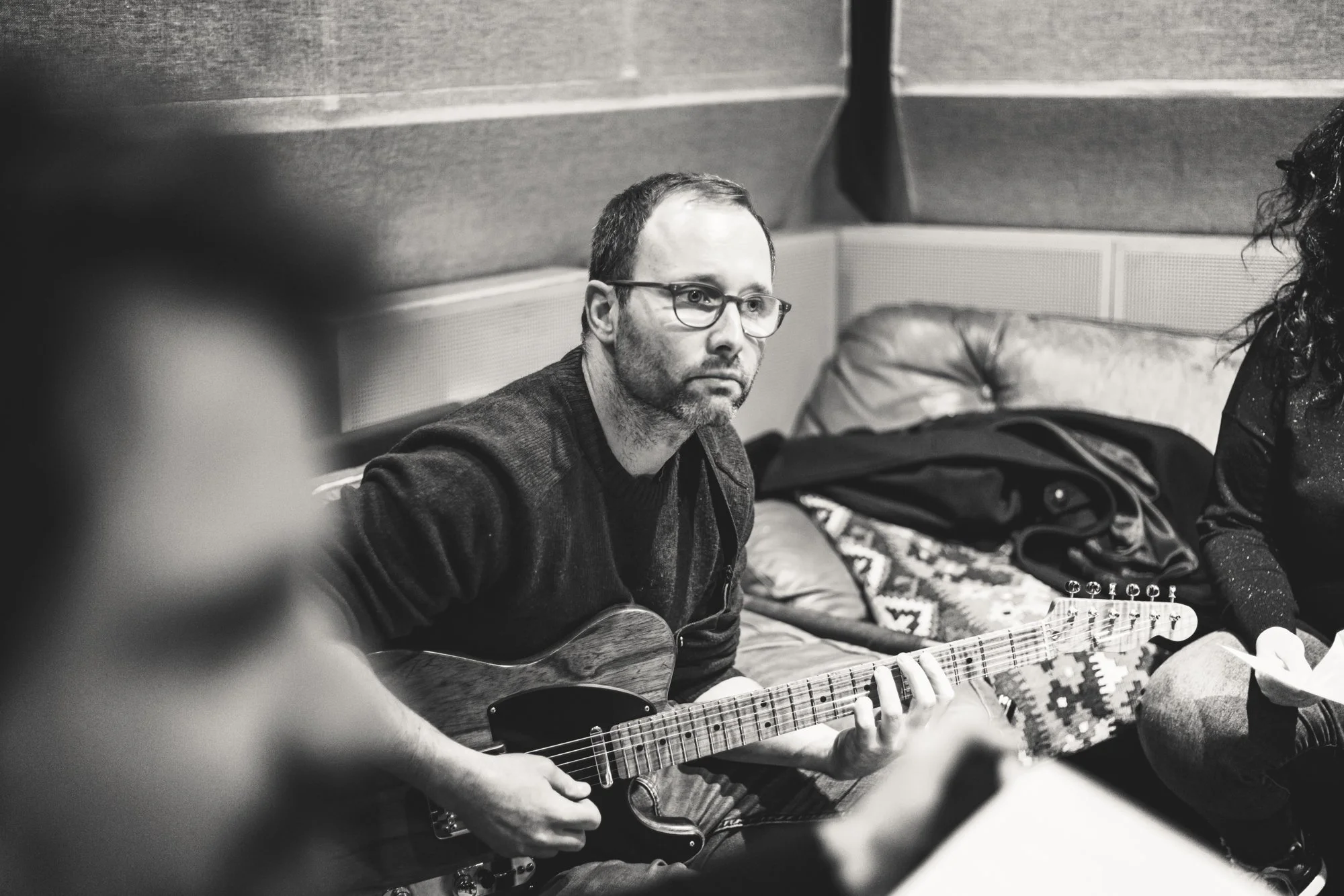 A man with glasses and a beard playing an electric guitar in a casual indoor setting, with a woman partially visible sitting nearby.