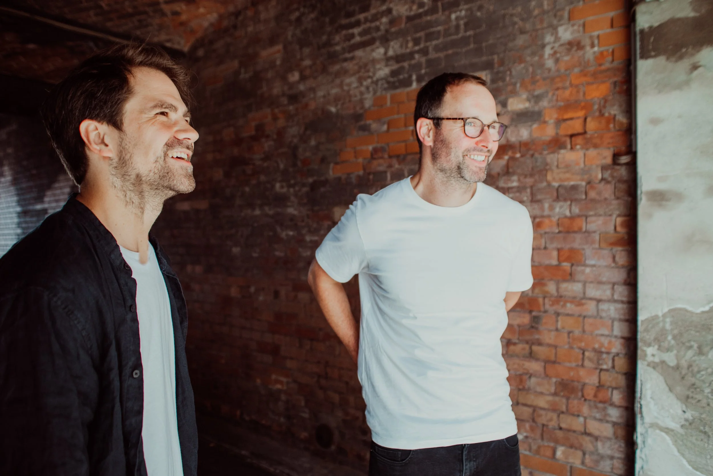 Two men smiling and talking in front of a brick wall, one wearing a black shirt and the other wearing a white T-shirt and glasses.