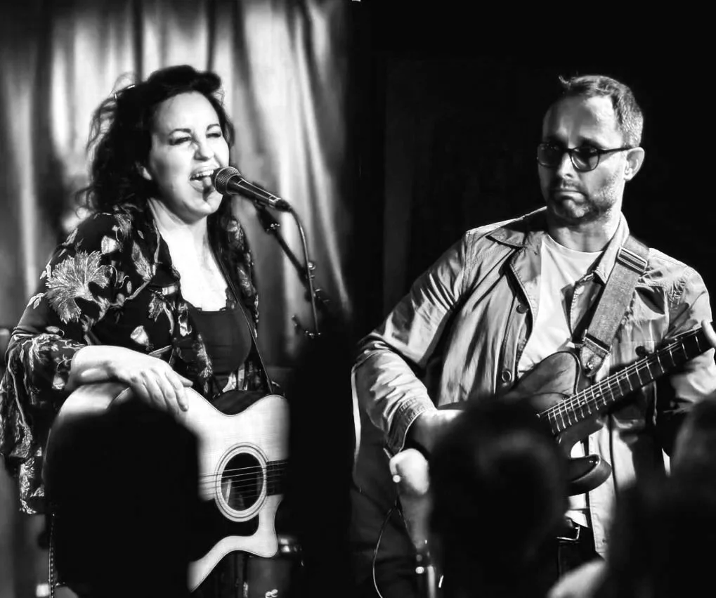 A woman singing and playing an acoustic guitar on stage, with a man playing an electric guitar beside her, both performing in front of an audience in a dimly lit venue.