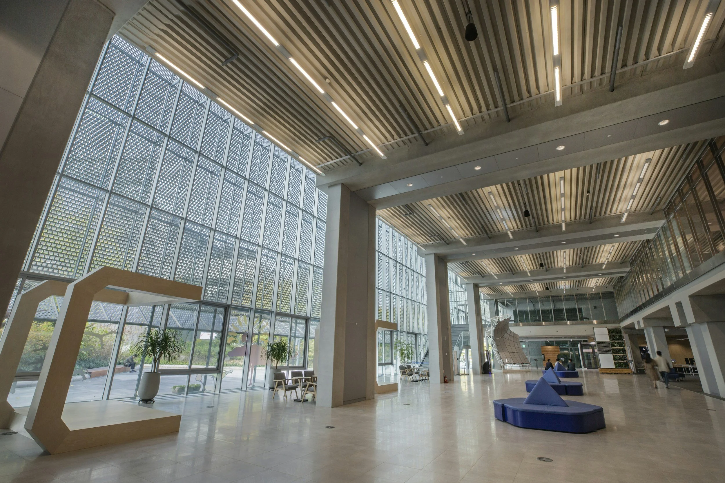 Modern interior space with large glass windows, wooden ceiling, and seating area with blue chairs and potted plants.