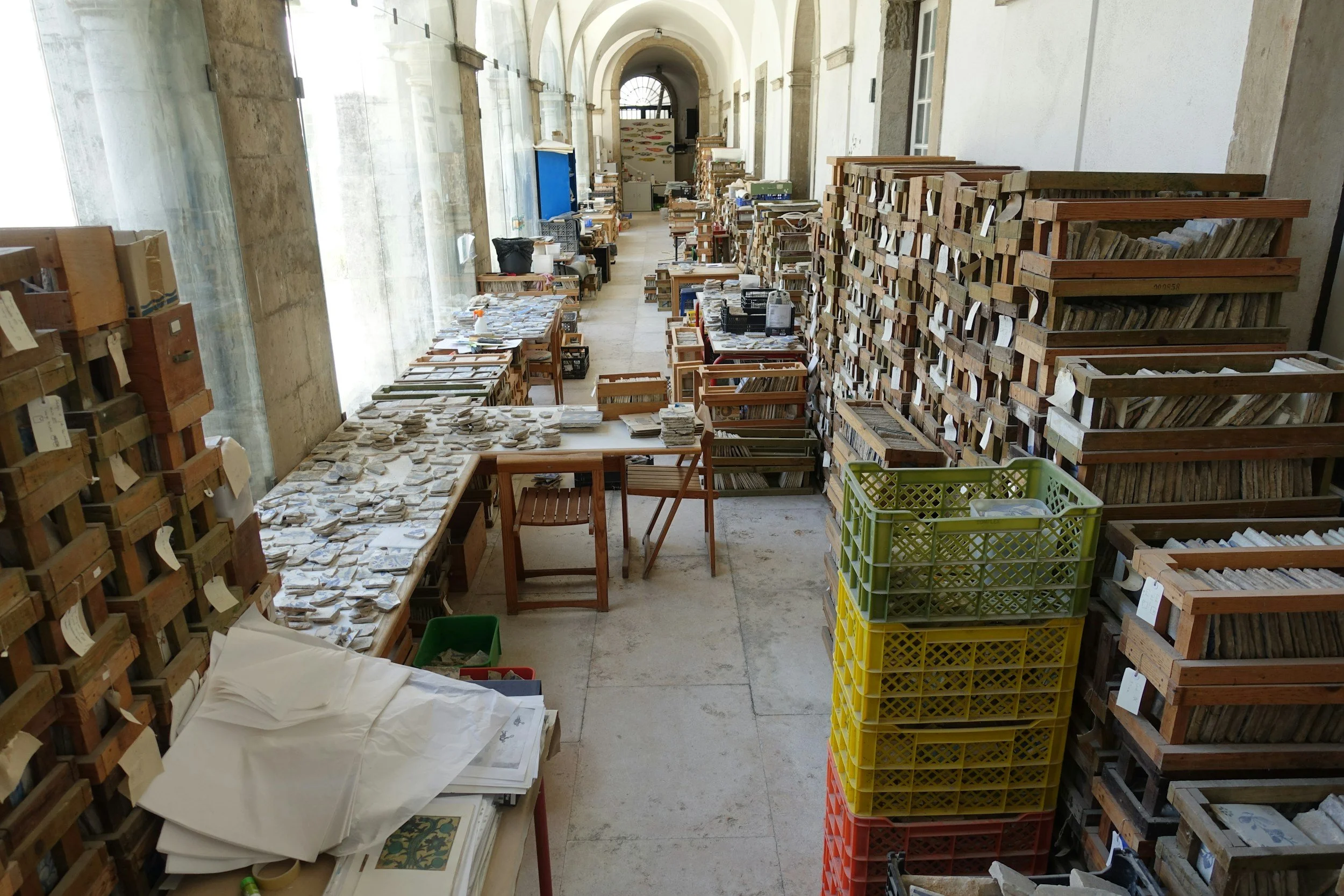 A long room with shelves filled with books and small artifacts, organized in wooden crates and stacks, with a few tables holding documents, in a historic building with high arched ceilings and large windows.