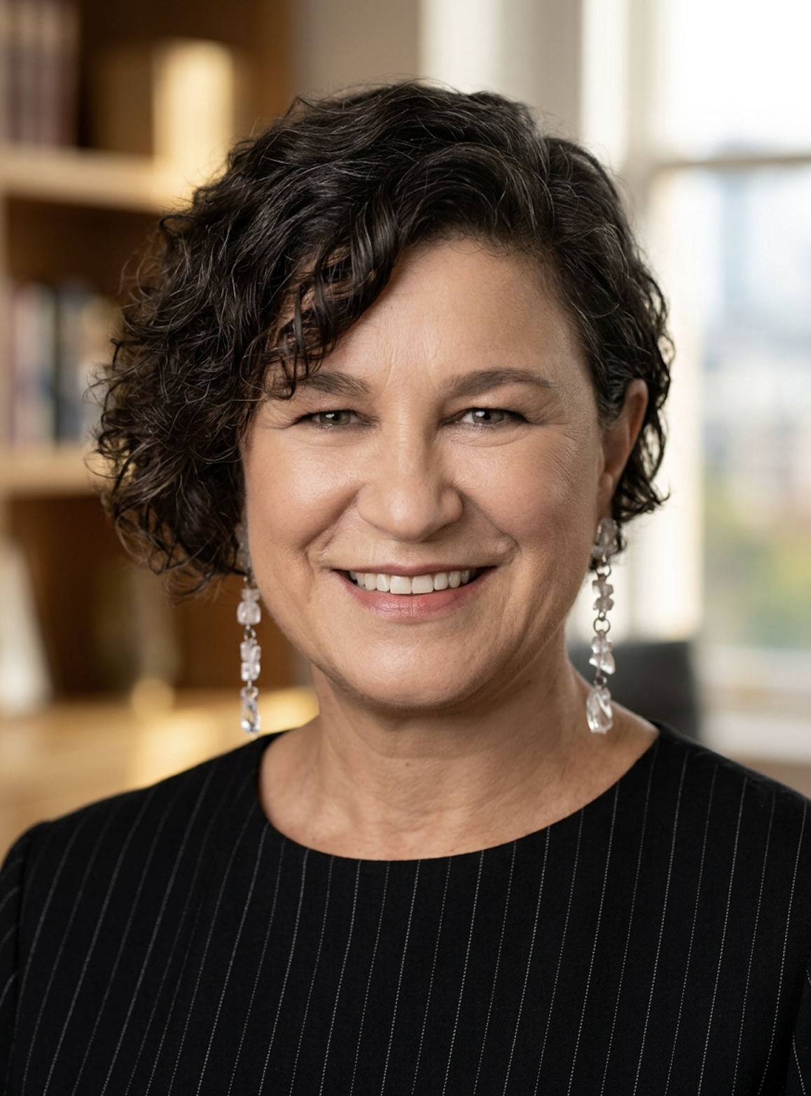 A middle-aged woman with short, curly dark hair, smiling, wearing dangling earrings and a black pinstripe top. She is in an office setting with bookshelves in the background.