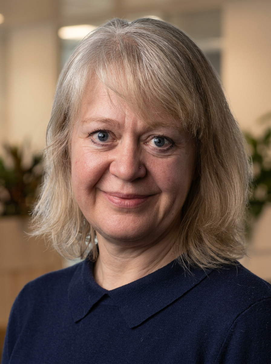 A woman with shoulder-length blond hair and blue eyes wearing a navy blue top, smiling in an indoor setting with blurred background.