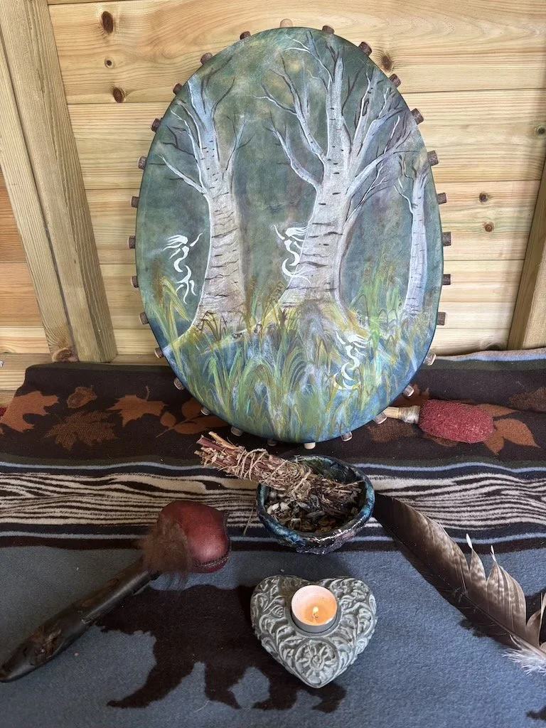 Decorative display featuring a painted oval wooden shield or plaque with two trees and white branches, a small bowl with a bundle of sage, a lit candle in a heart-shaped holder, a red stone, a large feather, a medallion, and a wooden stick with a red stone attached, all on a dark cloth with a printed patterned runner.