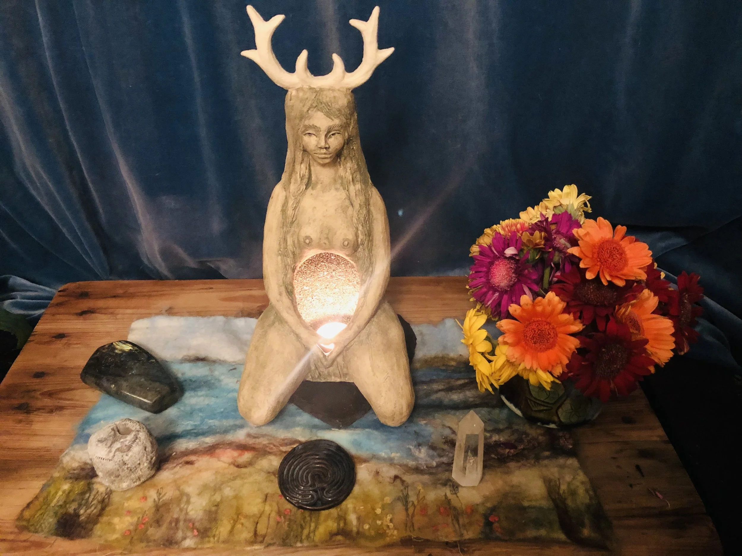 A decorative wooden sculpture of a woman with a face, mounted on a piece of wood with antlers on top, surrounded by colorful flowers and stones on a wooden surface with a dark blue backdrop.