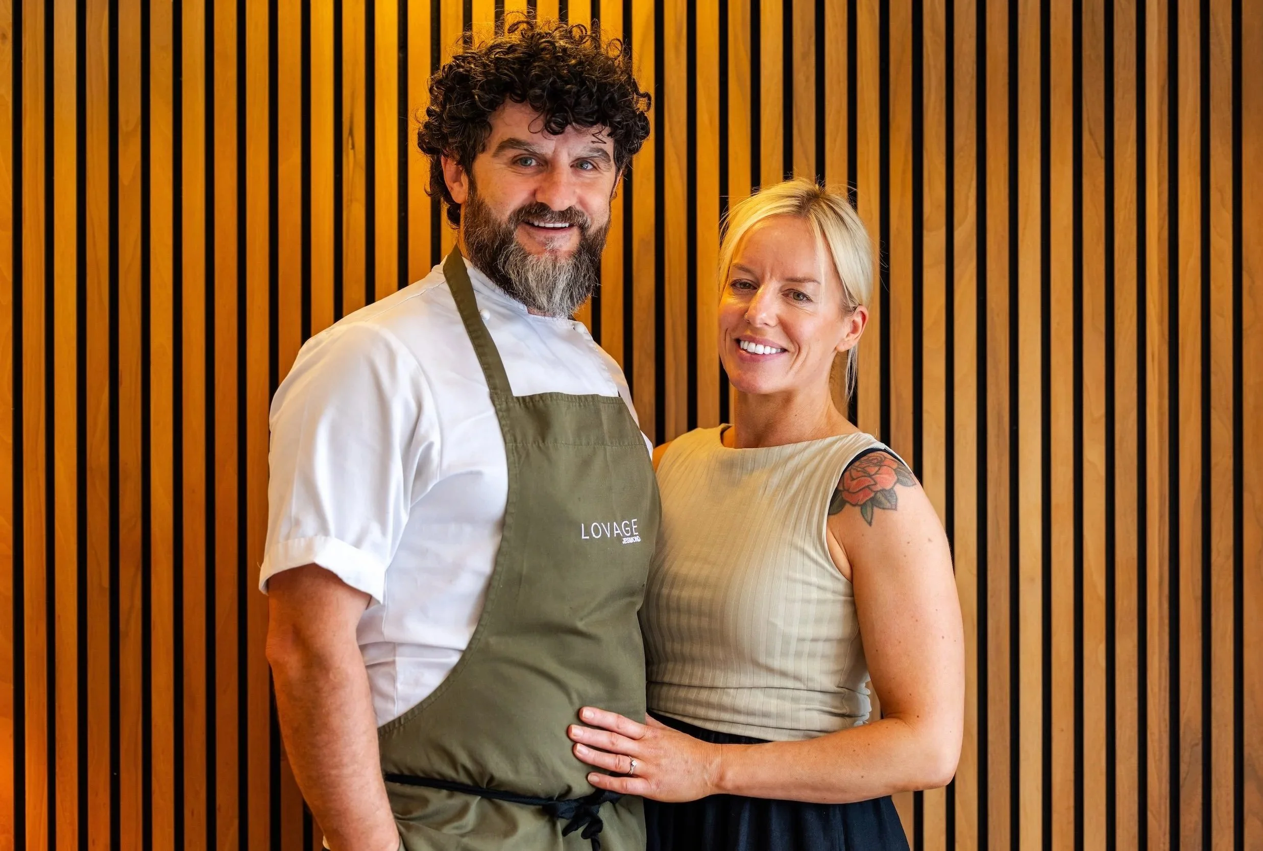 A man with curly dark hair and a beard, wearing a chef's coat and apron, standing next to a woman with blonde hair and a tattoo on her right arm, posing in front of a wooden slat wall.