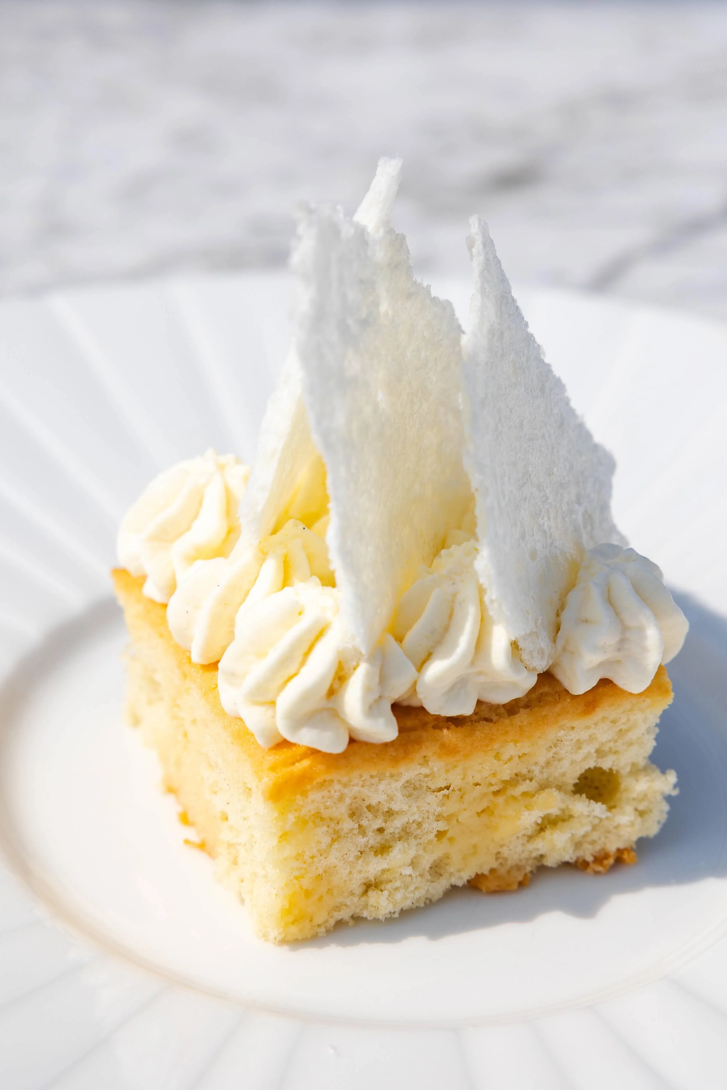A slice of vanilla cake topped with whipped cream and white chocolate shavings on a white plate.