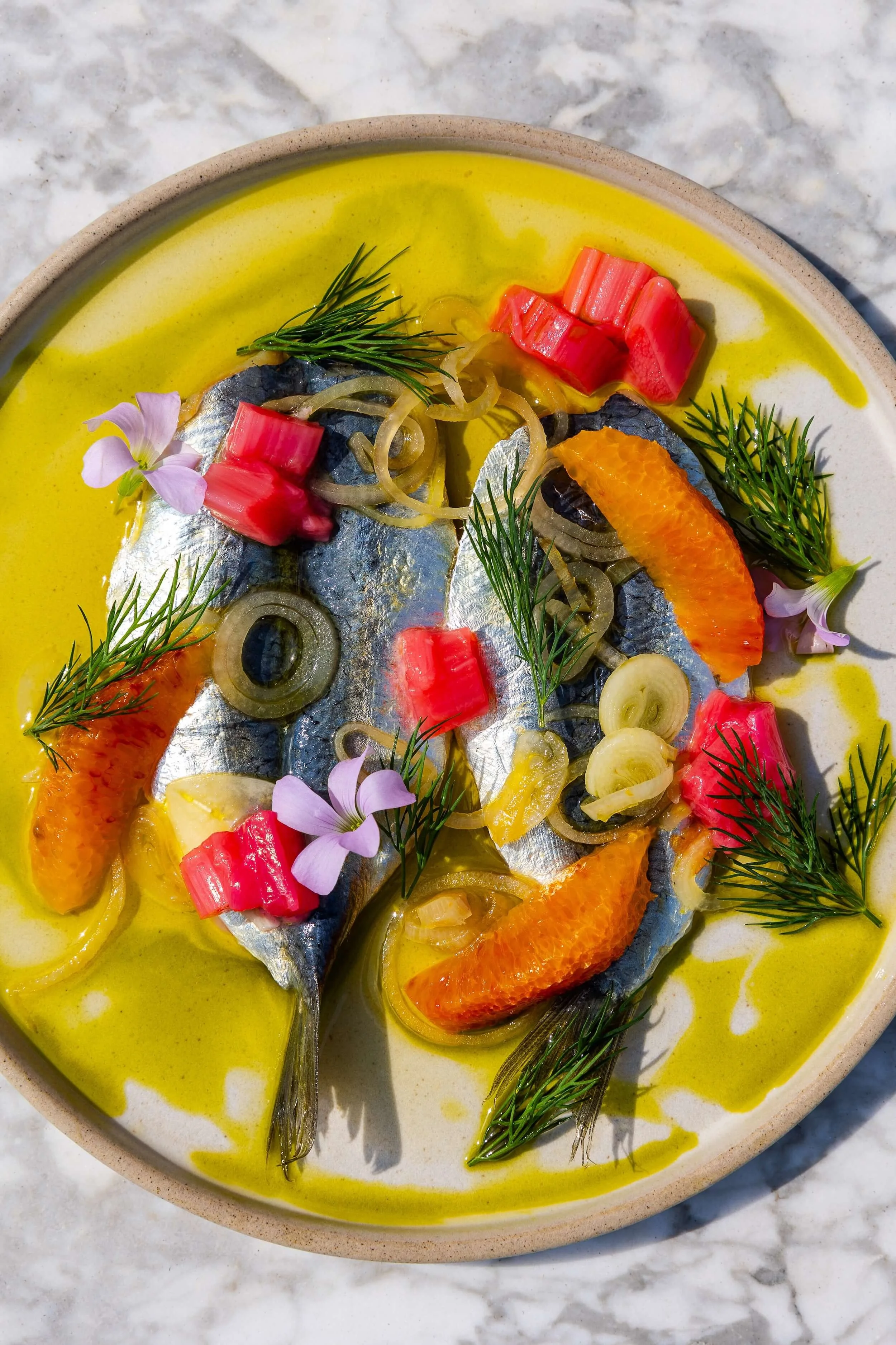 Dorado fish fillets topped with crab meat, sliced onions, orange slices, fresh dill, and edible flowers, served in a creamy yellow sauce.