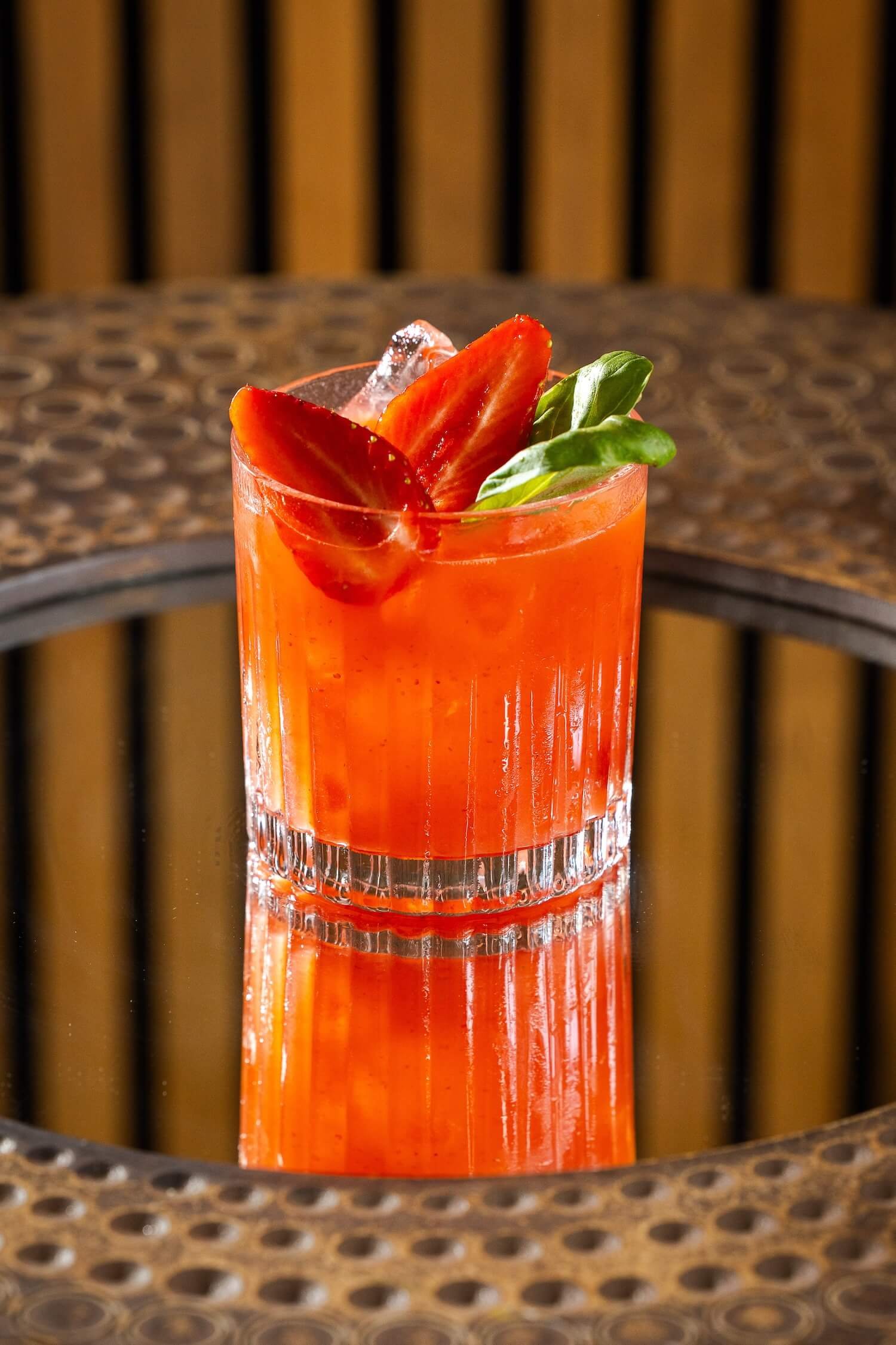 A glass of red fruit juice garnished with sliced strawberries and basil leaves, placed on a reflective surface.