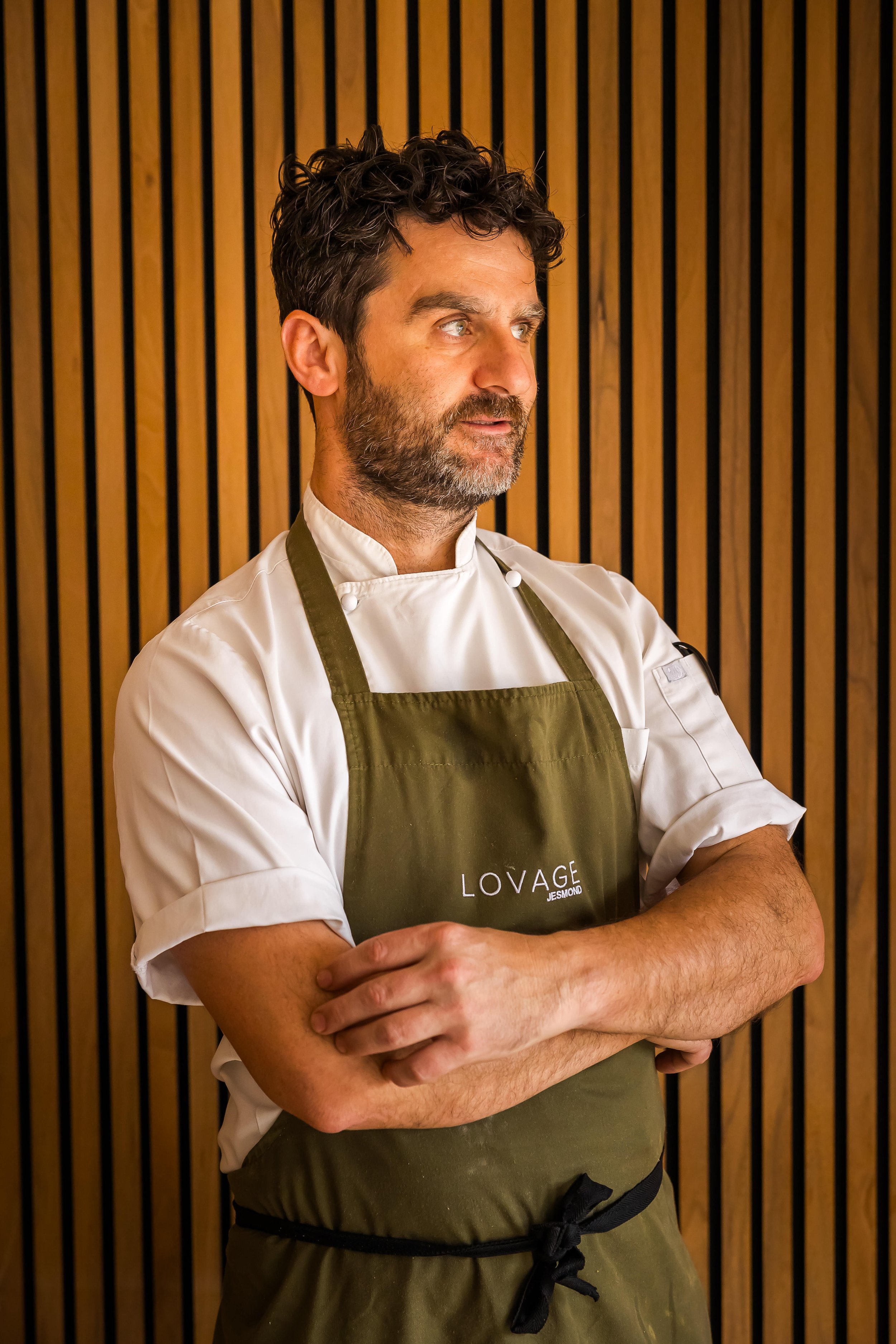 Chef Patron Kleo in a white chef's coat and olive green apron with the text 'LOVAGE JEMOND' on it, standing with arms crossed in front of a wooden slat wall.