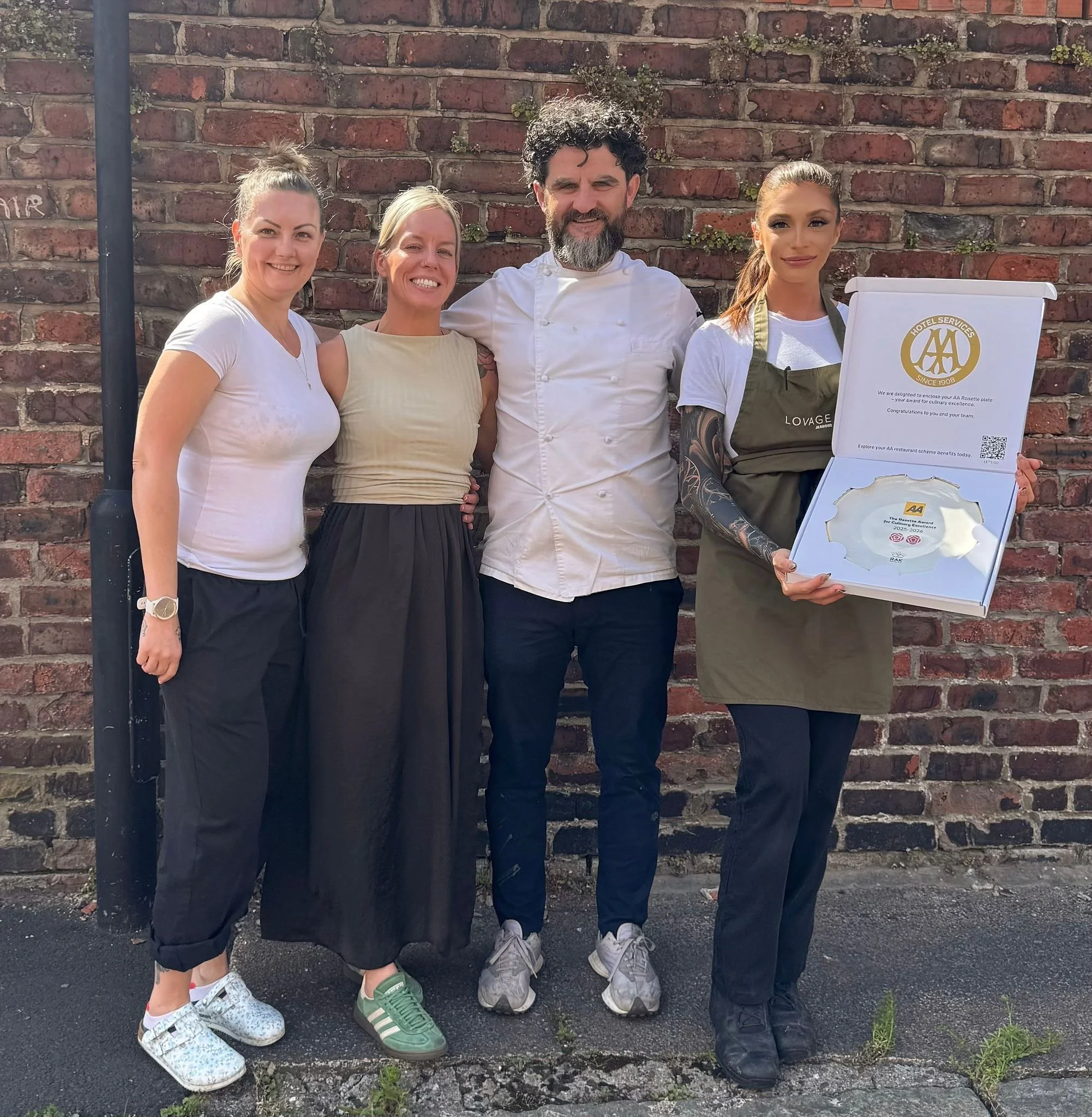 Four women and one chef standing together in front of a brick wall, smiling at the camera. One of the women is holding a sign or menu, and the group appears to be celebrating or at an outdoor event.