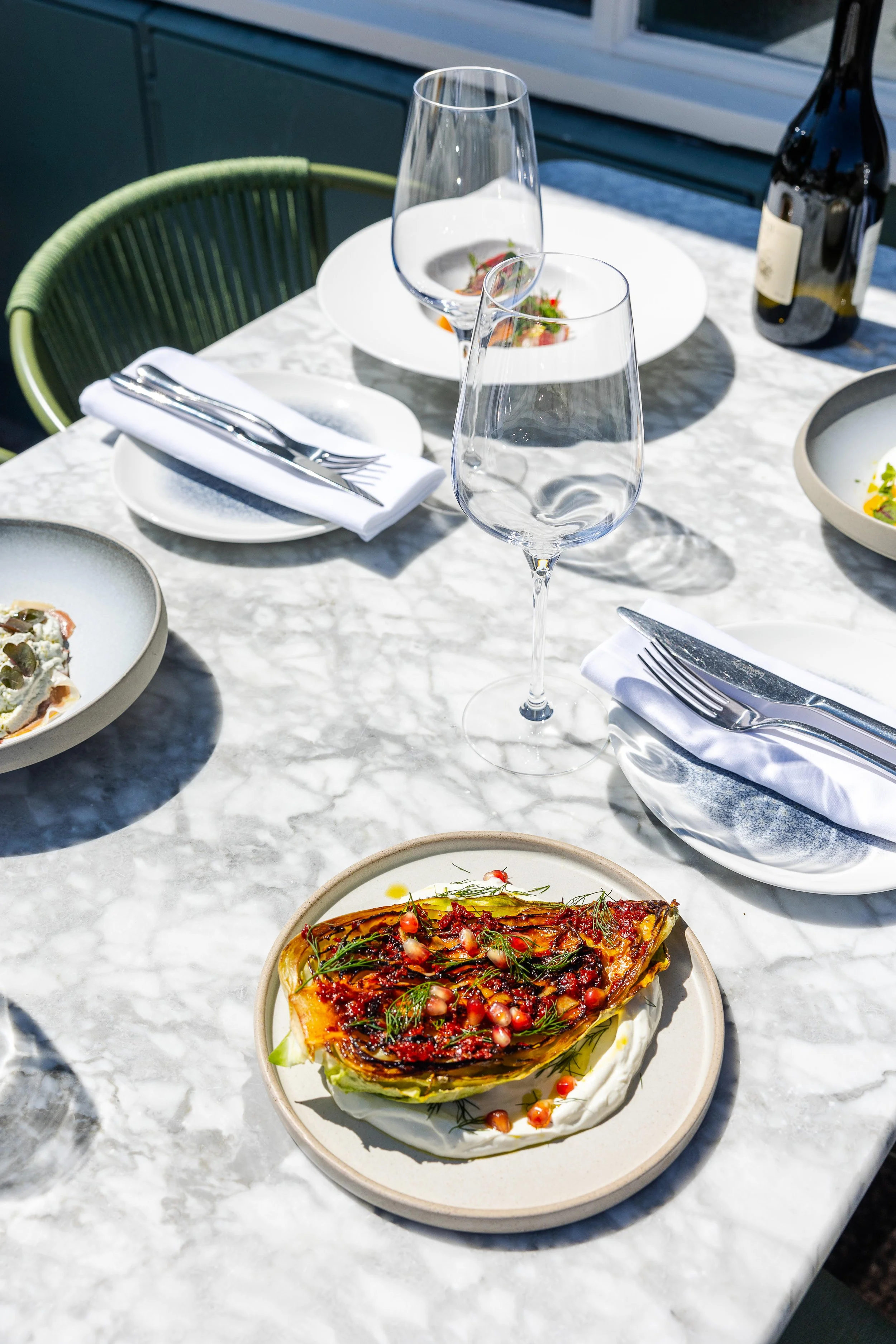 A table set for dining with a mozzarella and tomato appetizer, empty wine glasses, a napkin with silverware, a wine bottle, and a food plate with salad, all on a white marble surface.