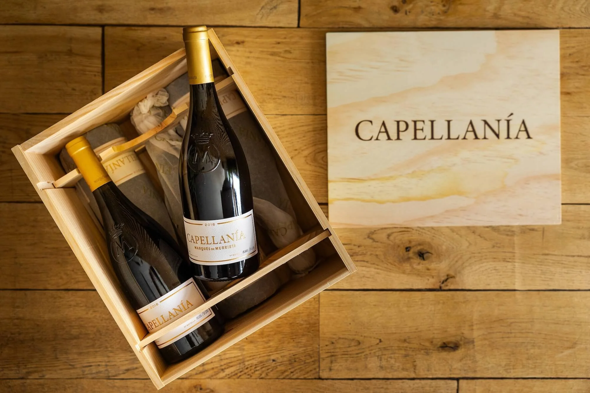 A wooden box containing several bottles of Capellanía wine with gold foil caps. The bottles are arranged inside the box, which is open. Next to the box is a wooden plaque or sign with the words "CAPELLANÍA" printed on it, placed on a wooden floor.