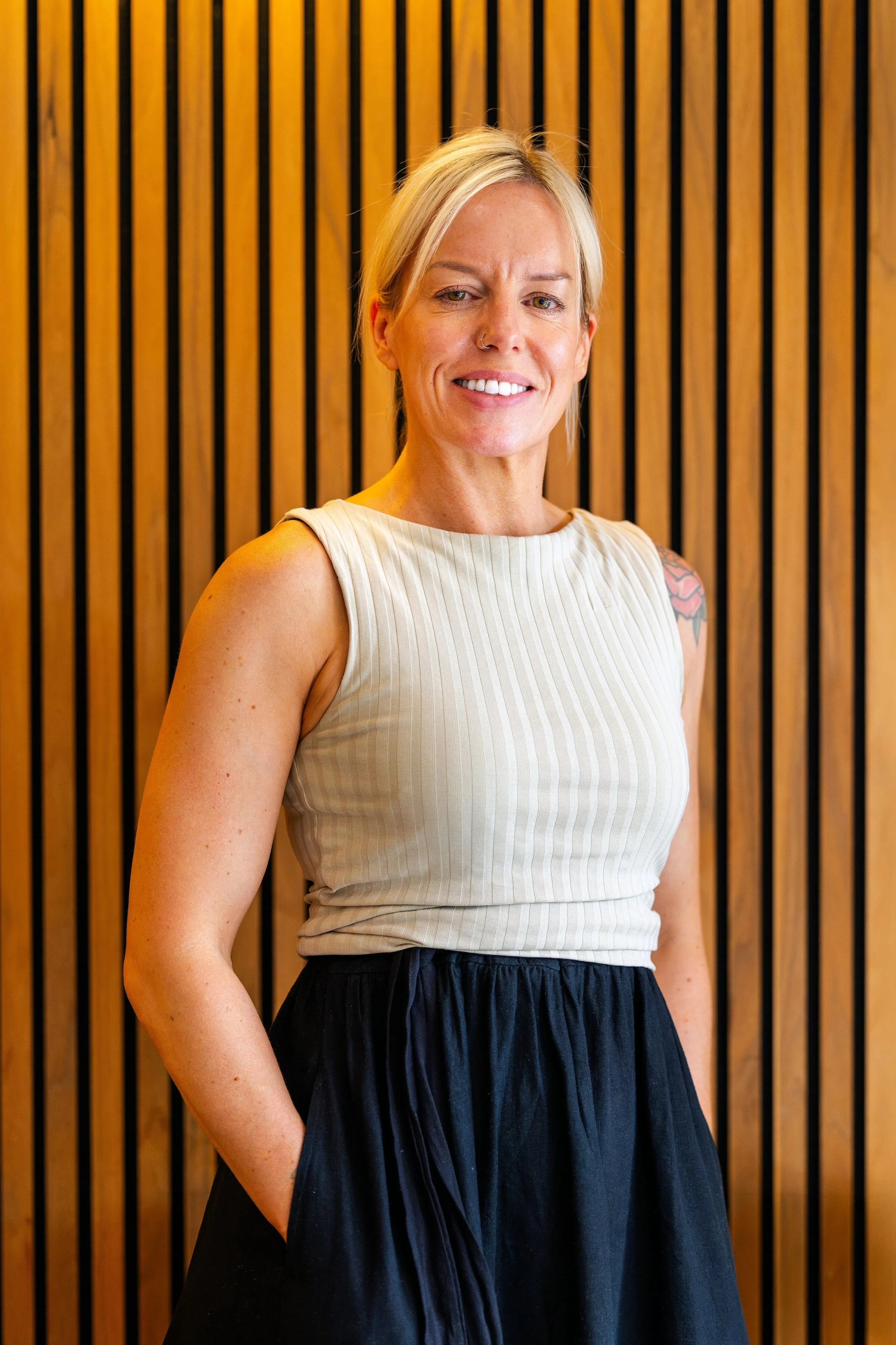 A smiling woman with blonde hair and a nose ring, wearing a sleeveless beige top and a black skirt, standing in front of a wooden slat wall.