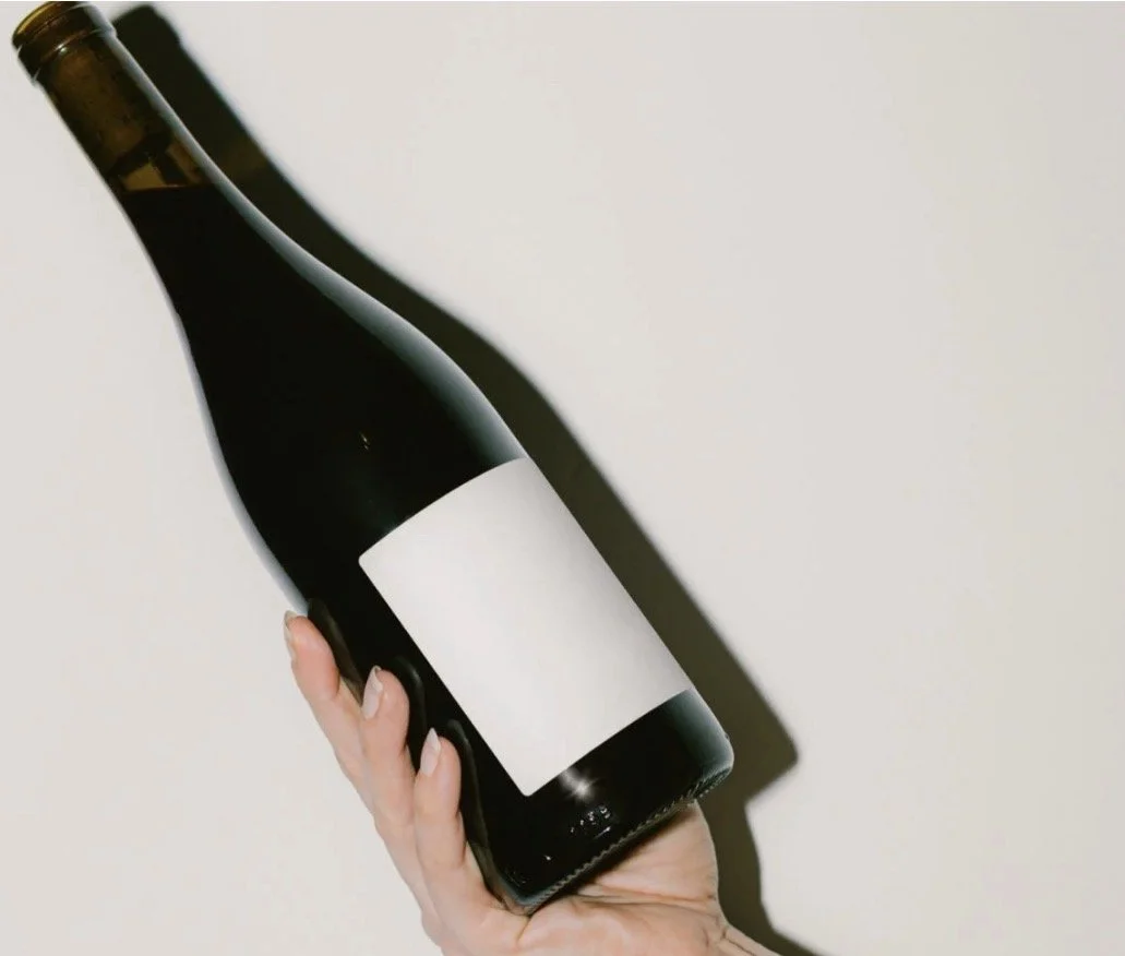 Hand holding a dark glass wine bottle with a blank white label against a plain background.