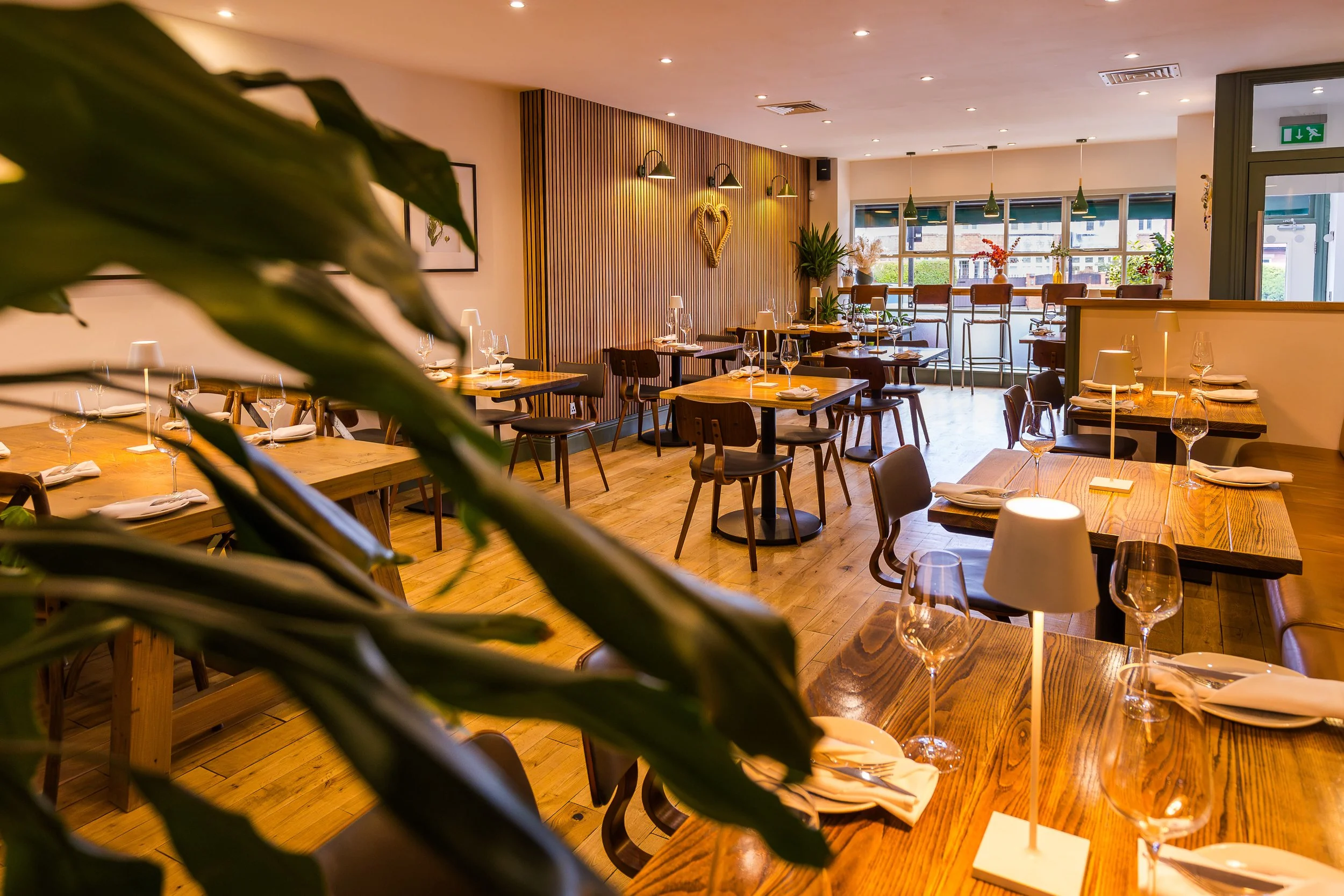 A cozy restaurant with wooden tables set with white napkins and wine glasses, decorated with warm lighting, green wall lamps, and plants, featuring large windows and wall art.
