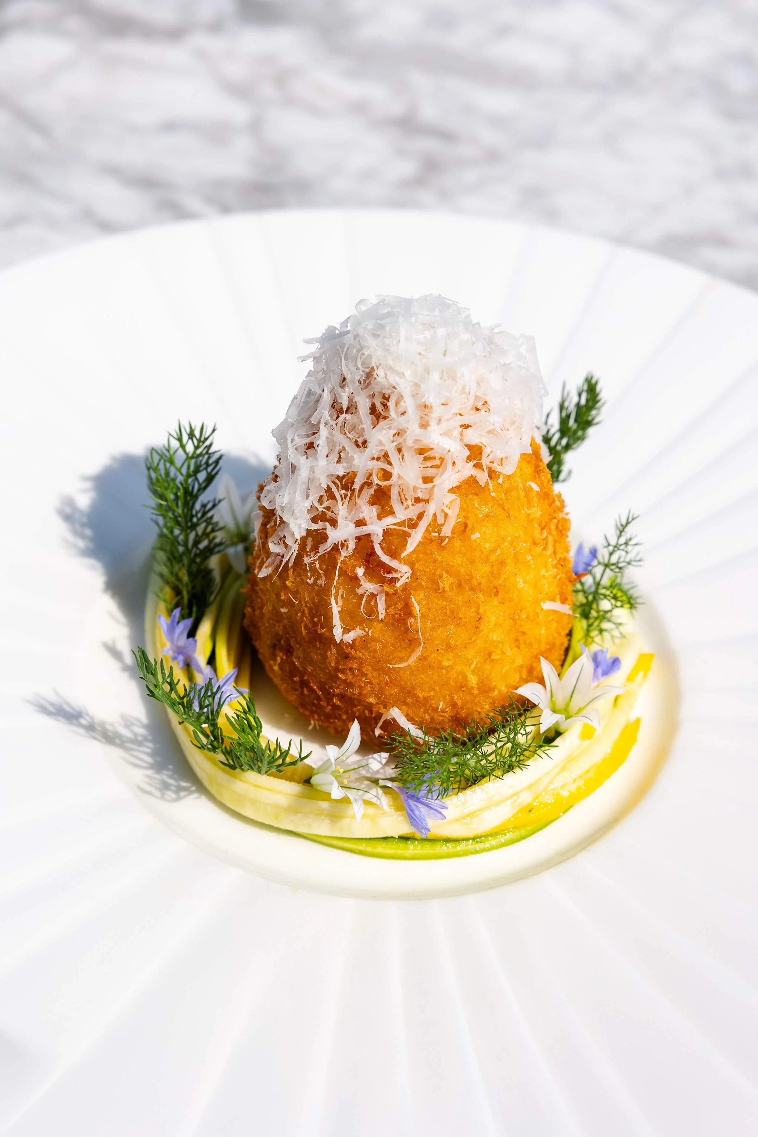 Breaded and fried food topped with shredded cheese, garnished with small green herbs and purple and white edible flowers, served on a white plate.