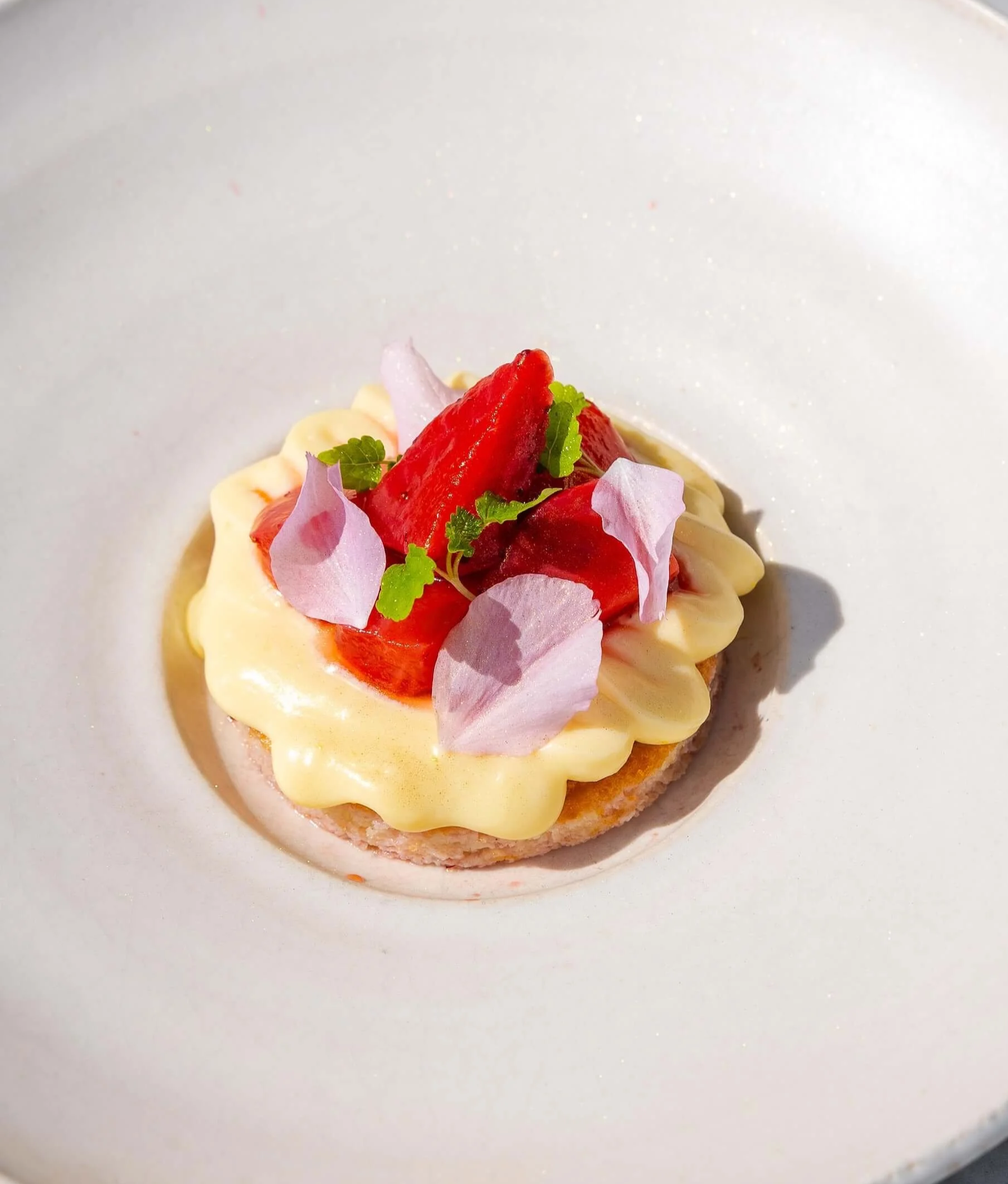 Dessert cake topped with strawberries, edible flower petals, and green herbs on a white plate.
