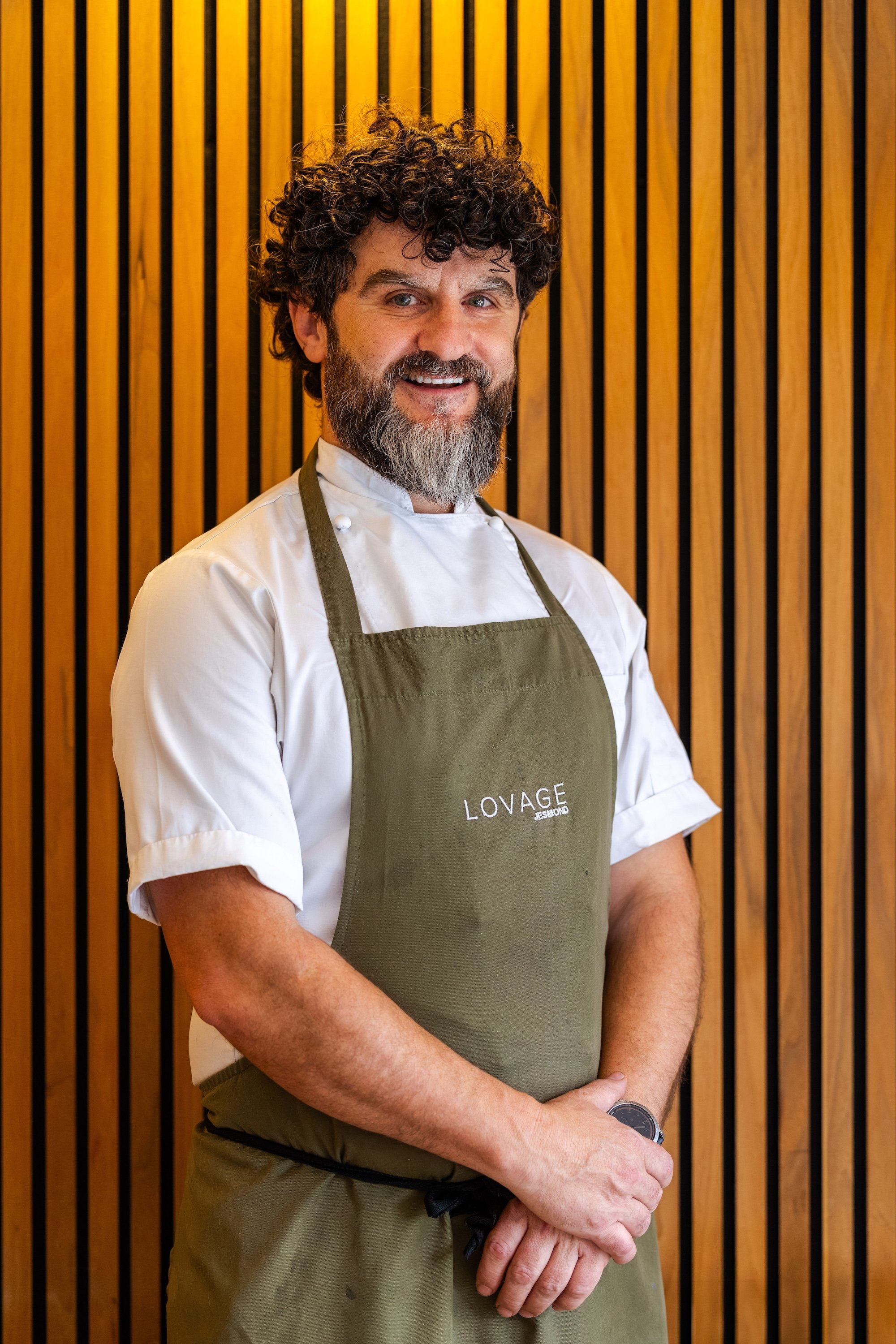 A man with curly hair and a beard smiling, wearing a white shirt and a green apron with the word 'LOVAGE' on it, standing in front of a wooden slat background.