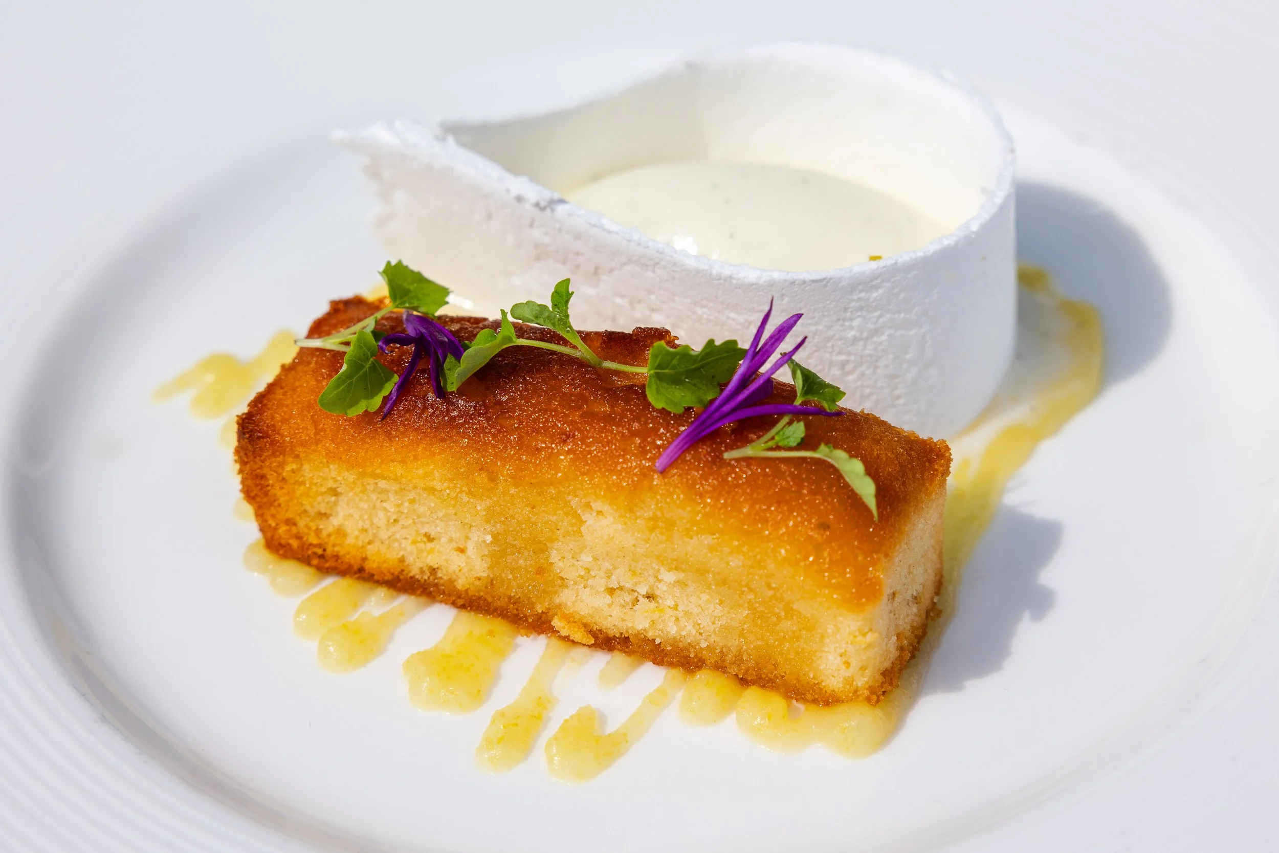 A slice of lemon-flavored cake topped with edible purple and green flowers, served with a dollop of cream or soft cheese on a white plate.