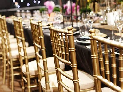 Gold chiavari chairs arranged around a banquet table with black tablecloths, set with glasses and floral centerpieces for an event or celebration.