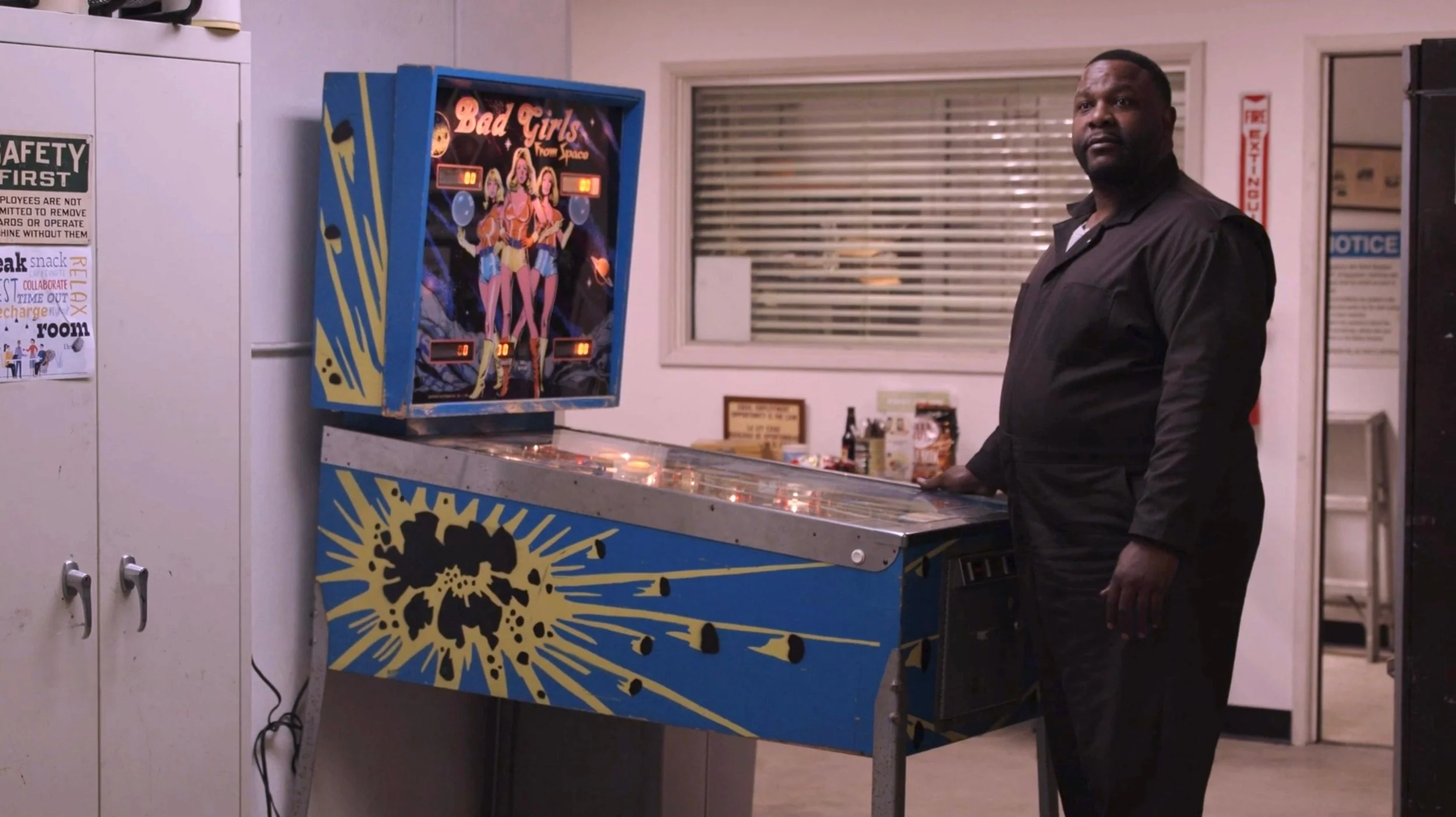 A man in a black shirt playing a pinball machine titled 'Bad Girls from Space' in an indoor setting. There is a cabinet with safety instructions and a window behind him.