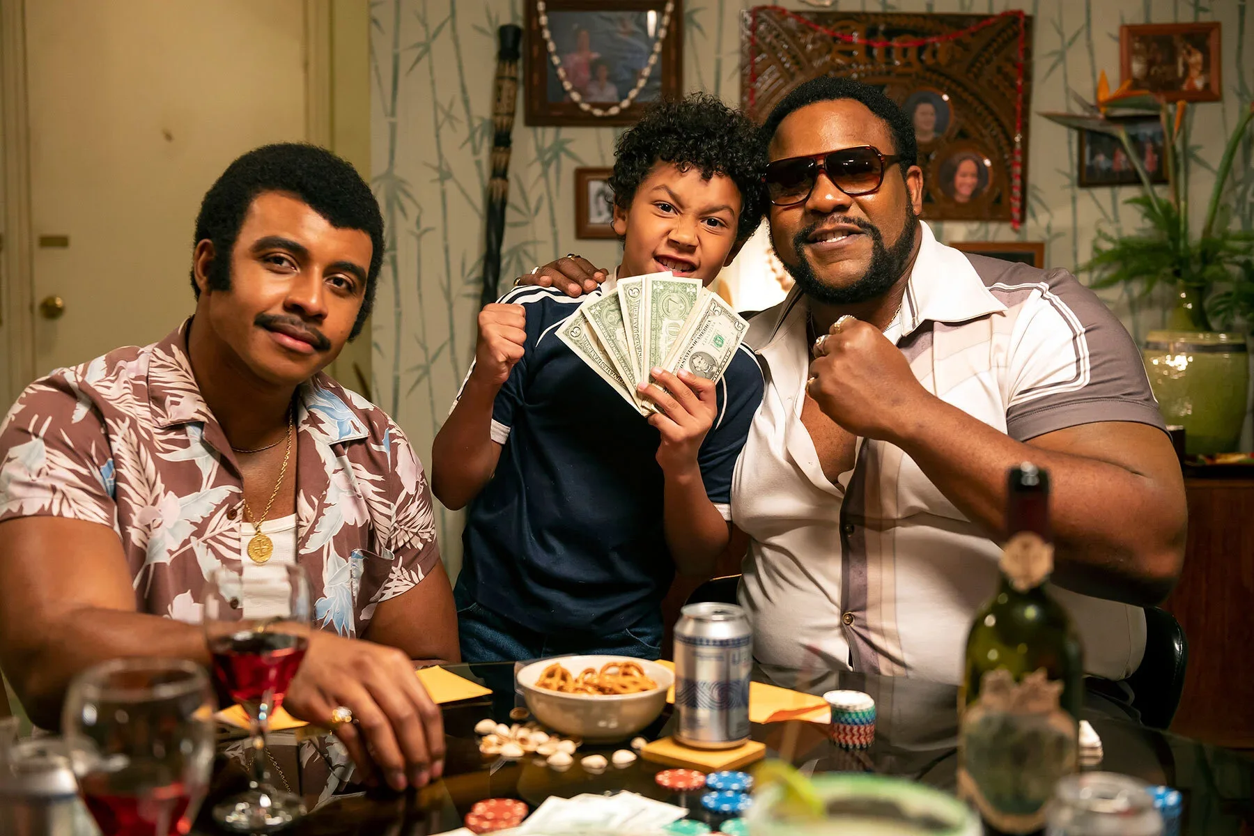 A group of four people celebrating at a dinner table with steaks, pasta, and drinks, one child holding cash, smiling and showing a rebellious expression, in a cozy, decorated living room.