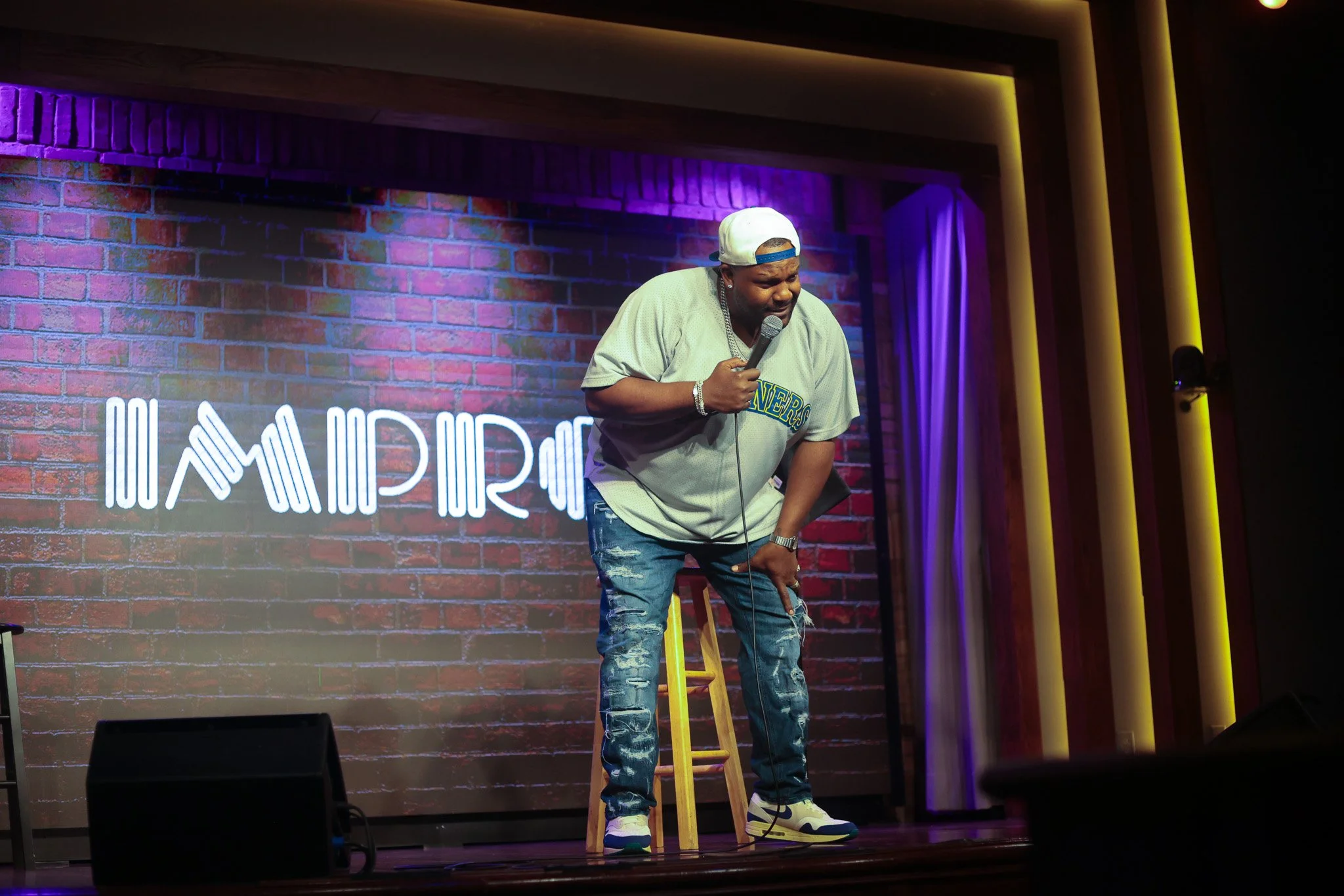 A comedian performing on stage with a microphone, wearing a white baseball cap backward, a beige T-shirt, and ripped jeans, standing next to a yellow stool. The stage background includes a brick wall with colorful lighting.