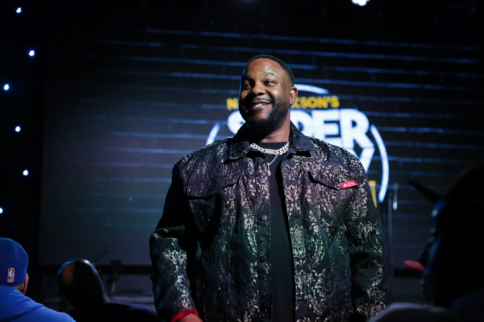 A man smiling wearing a patterned black jacket and jewelry, standing on a stage with a backdrop including the NBA logo and the words 'NBA All-Star Weekend'.