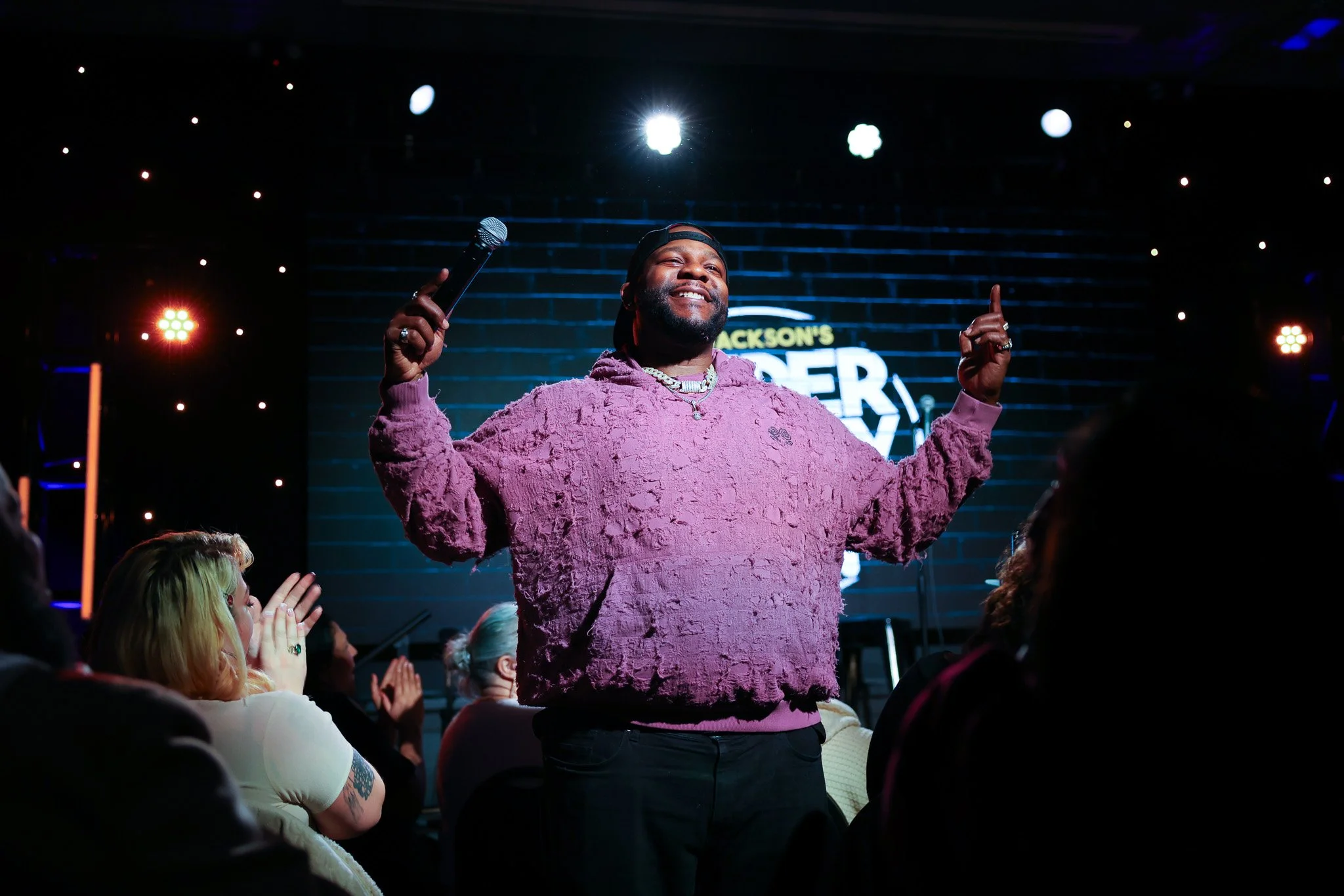 A man on stage holding a microphone in one hand, smiling, wearing a pink textured sweatshirt, baseball cap, and jewelry. Audience members are clapping and watching him, with a backdrop displaying the name 'Jackson's' and a neon marquee.
