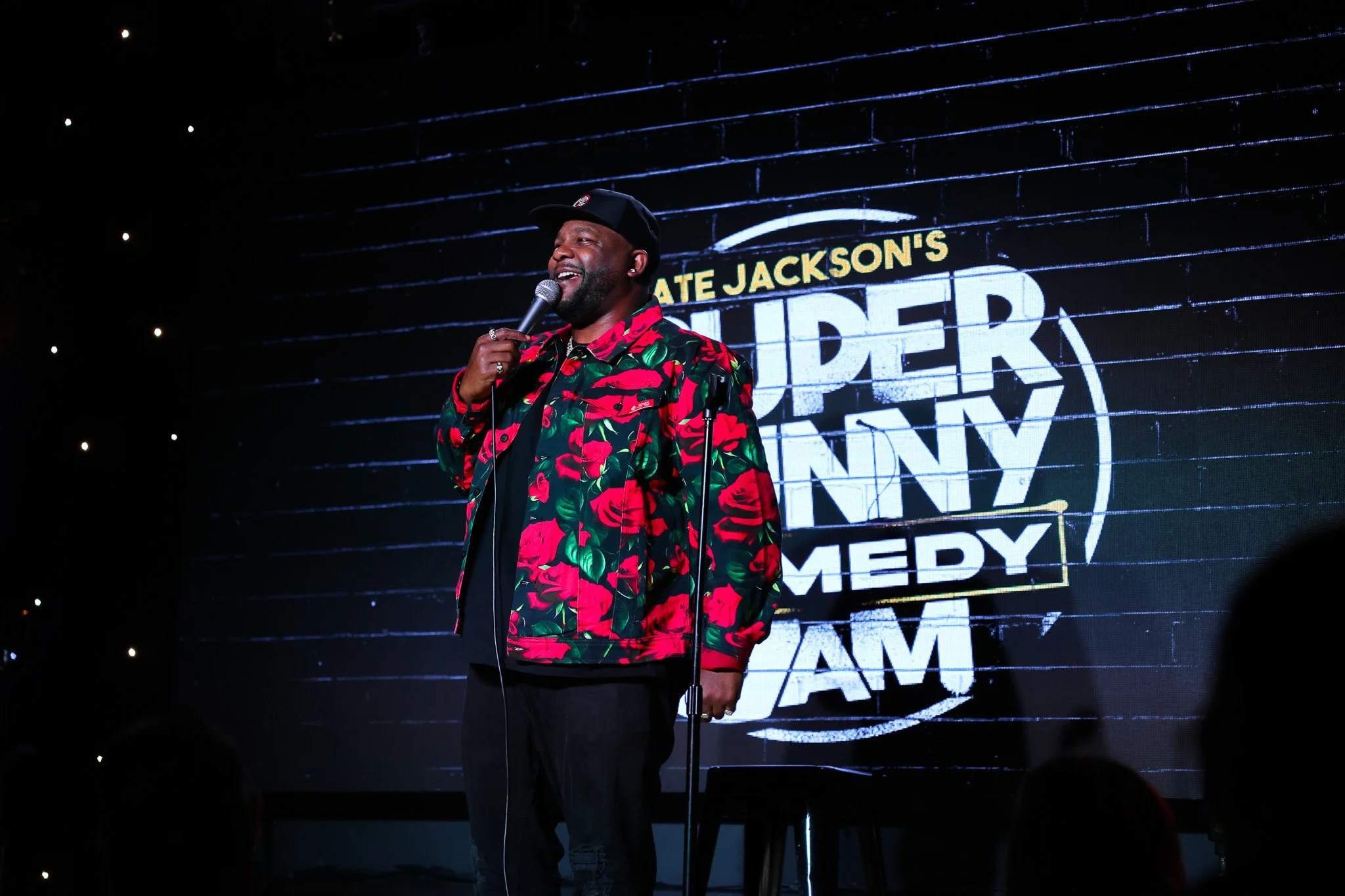 A man standing on stage speaking into a microphone, wearing a black cap and a red and green floral jacket, with a large screen behind him displaying text about comedy.