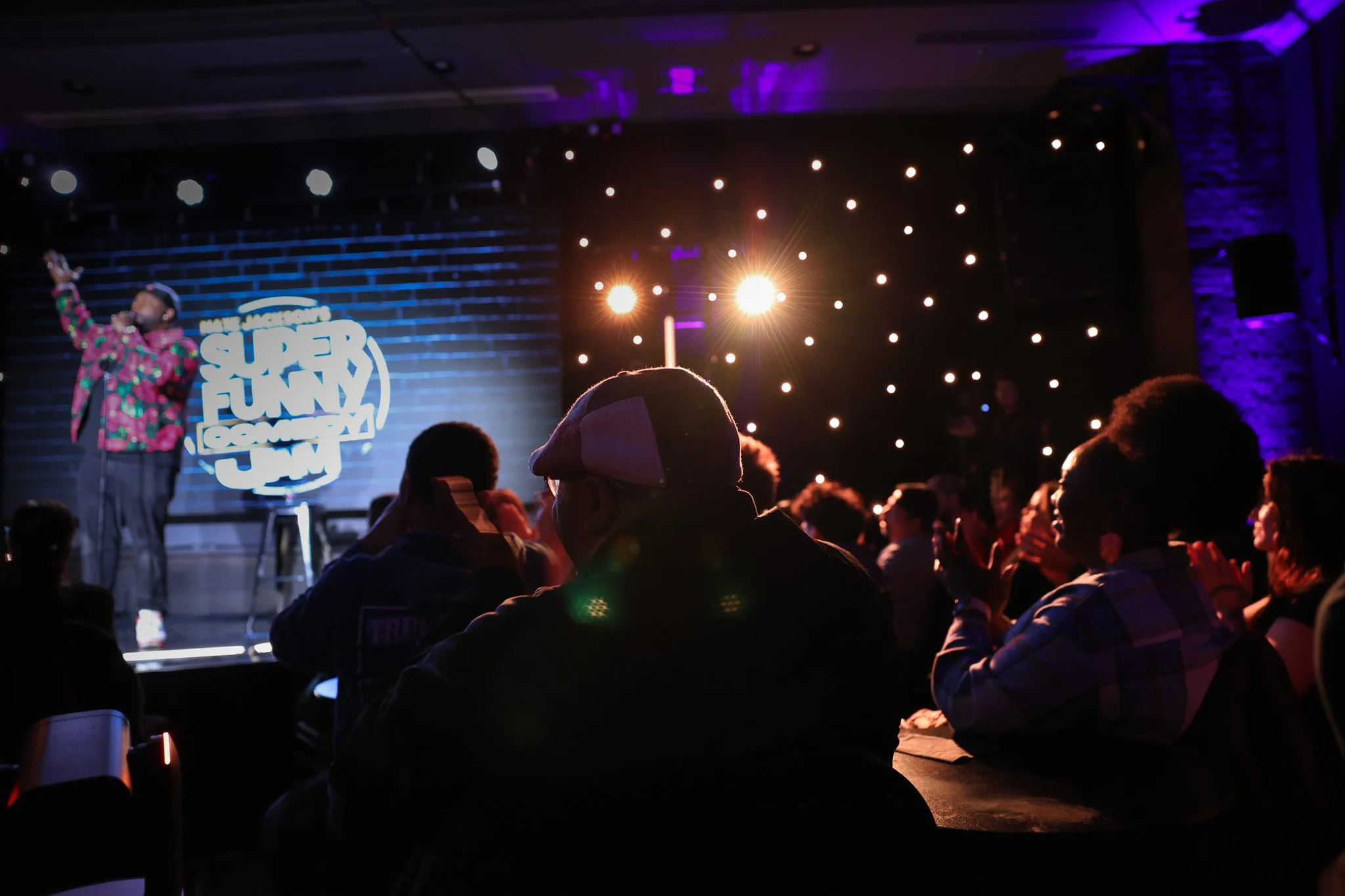 A performer on stage at a comedy show with a sign that reads 'Super Funny Comedy', surrounded by audience members seated in a dimly lit room with purple and blue lighting.