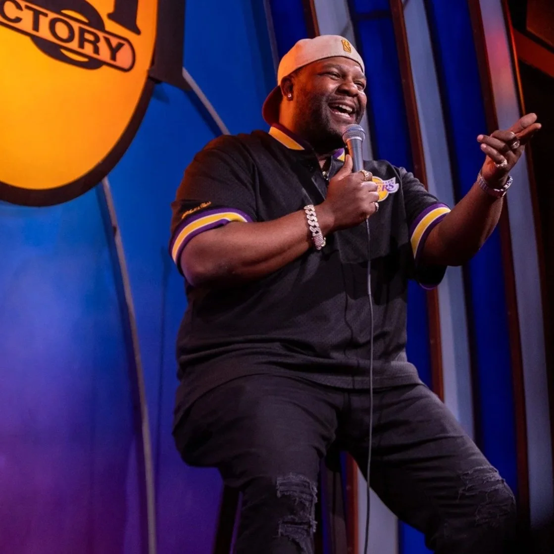 A man performing stand-up comedy on stage at a comedy club, holding a microphone, wearing a black Lakers sports jersey, a beige cap worn backwards, and jewelry, with a comedy club logo visible in the background.