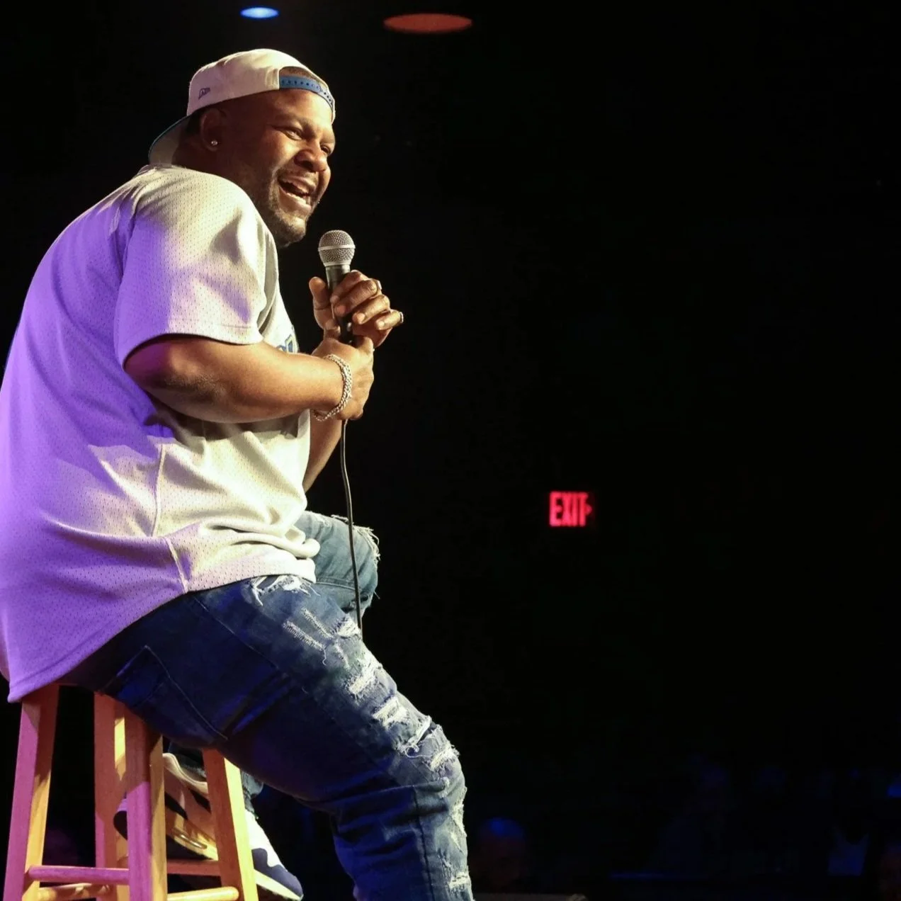 A man wearing a backwards cap, sports jersey, and ripped jeans sitting on a stool on stage, holding a microphone and smiling, with a dark background and a lit EXIT sign behind him.