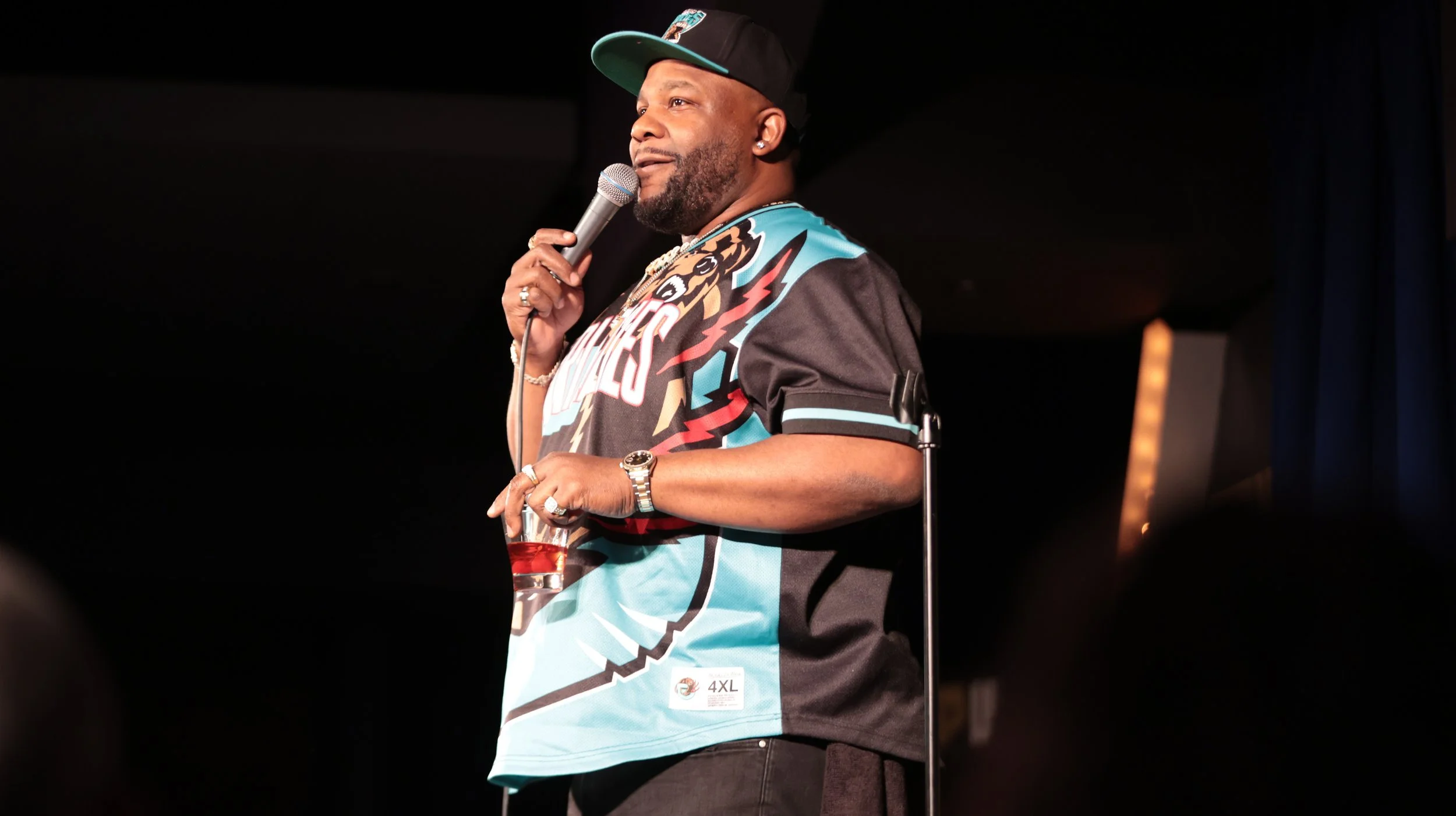 A man with a beard wearing a black and teal cap, a black, teal, and multicolor sports jersey, holding a microphone in one hand and a drink in the other, standing on stage against a dark background.