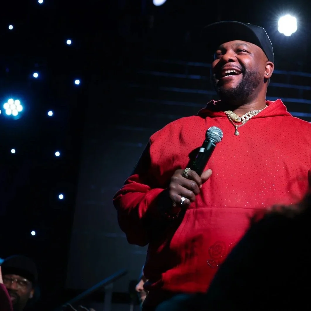 A man wearing a black cap and a red hoodie, smiling and holding a microphone, standing on stage with dark background and bright stage lights.