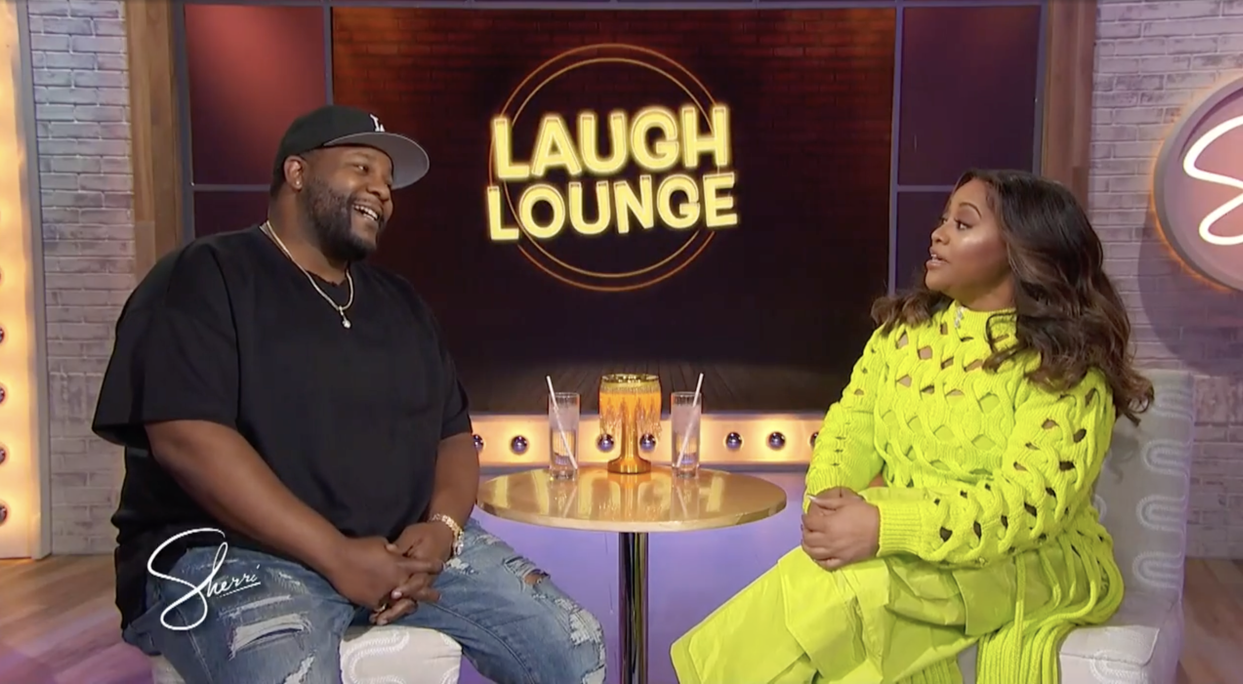 Two people, a man and a woman, sitting at a table and talking in a studio with a 'Laugh Lounge' sign in the background. The man is wearing a black T-shirt, a baseball cap, and distressed jeans, while the woman is dressed in a bright yellow outfit. There are two glasses of water and a decorative orange vase on the table.